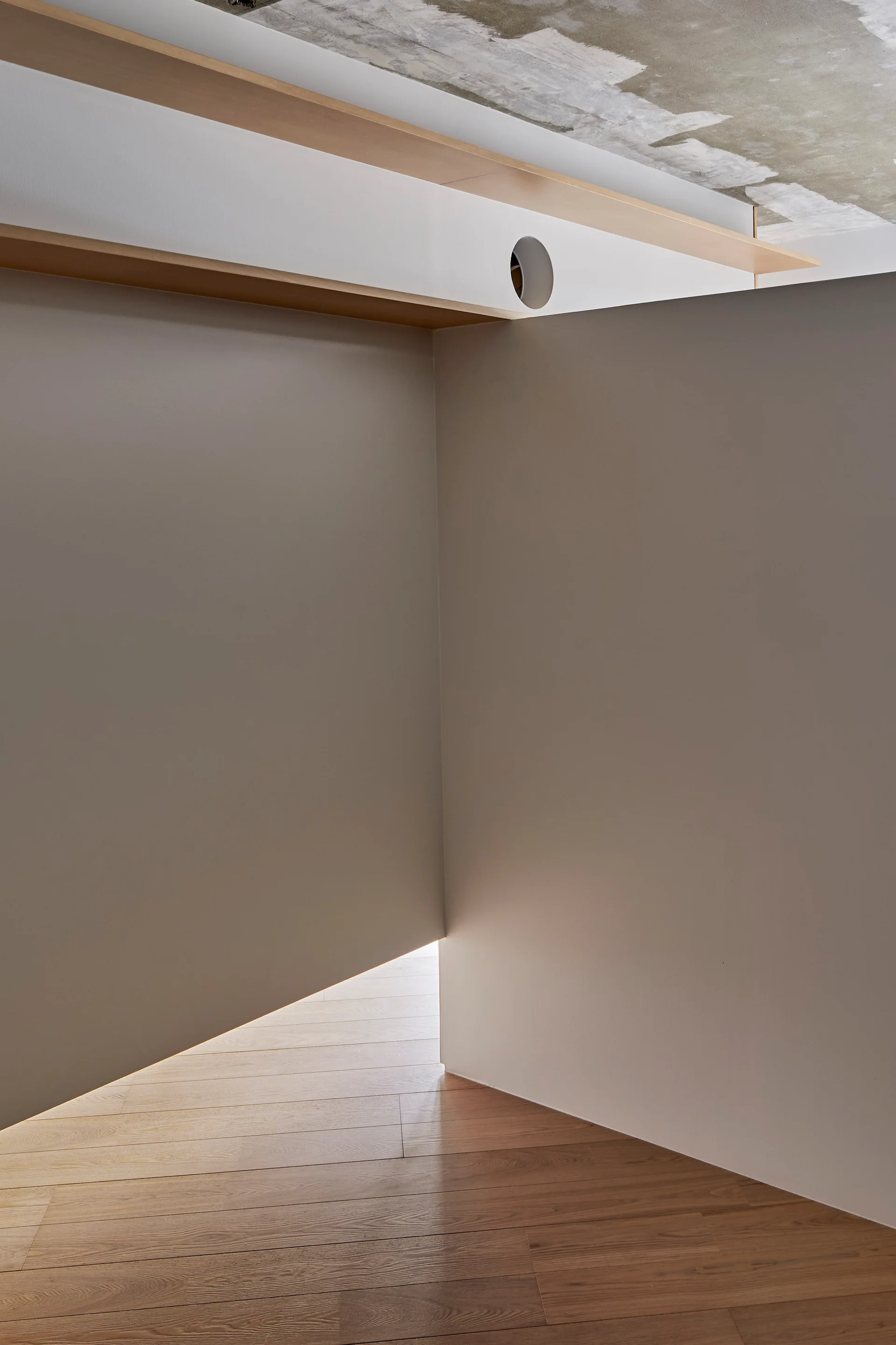 A minimalist grey corner wall with a gap at the floor showing warm light and wood planks, underneath a raw concrete ceiling.