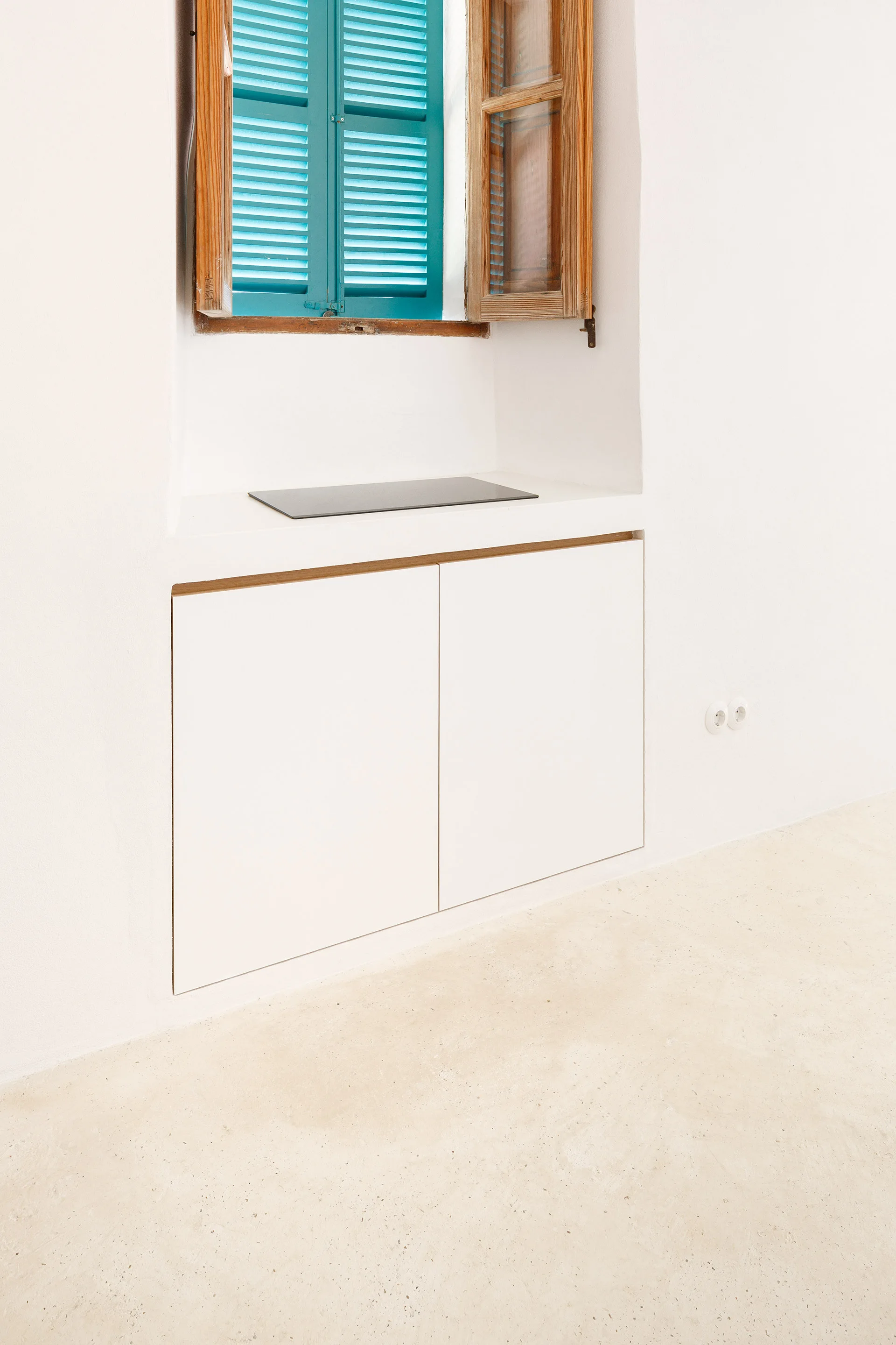 A minimalist white plaster wall features a recessed built-in cabinet beneath a window with bright blue wooden shutters and sand-toned stone flooring.