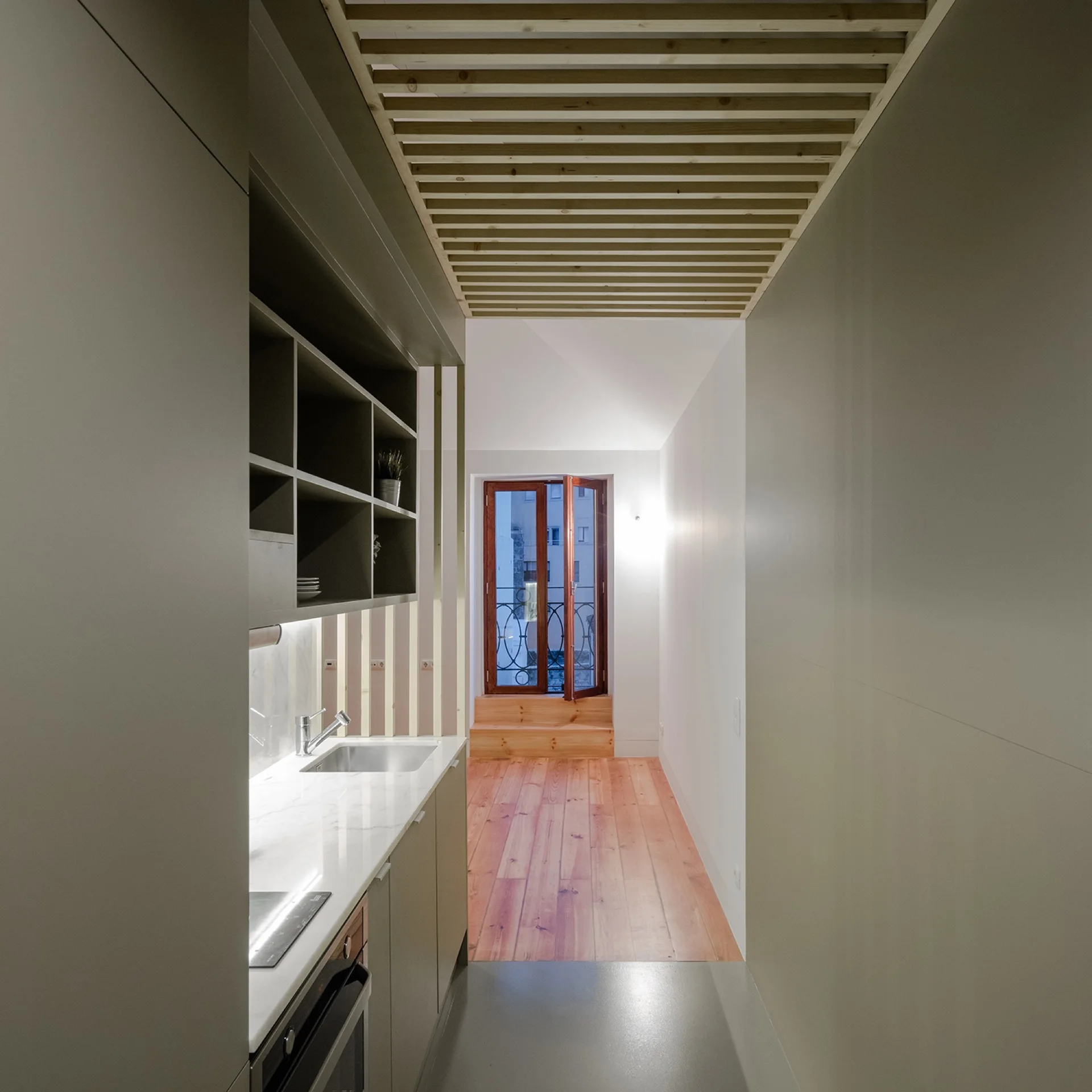 A narrow kitchen with gray cabinets and a wooden slatted ceiling leading to a hallway with wood plank flooring.
