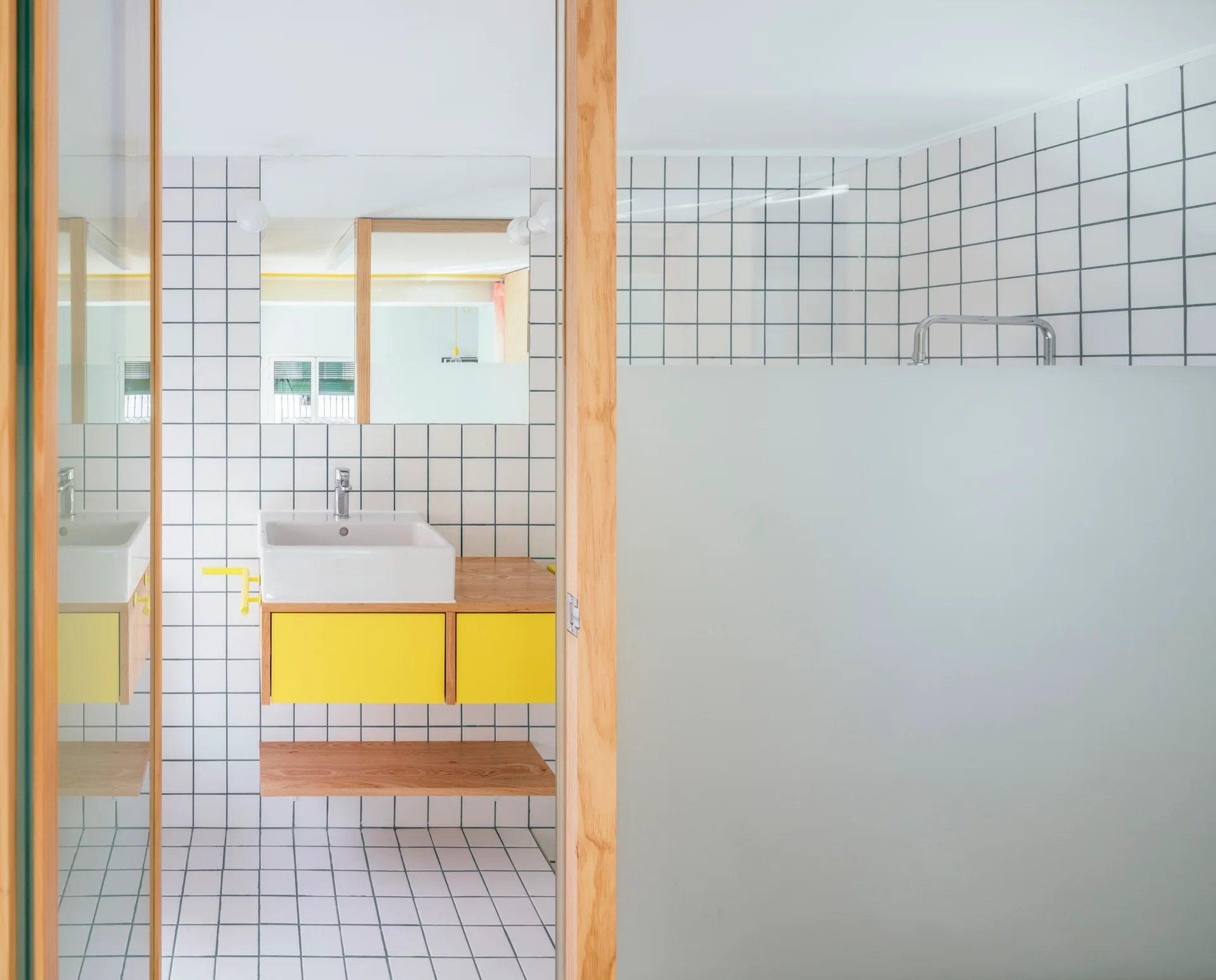 A modern bathroom features a floating yellow vanity with a white vessel sink against white grid-tiled walls and a frosted glass shower screen.