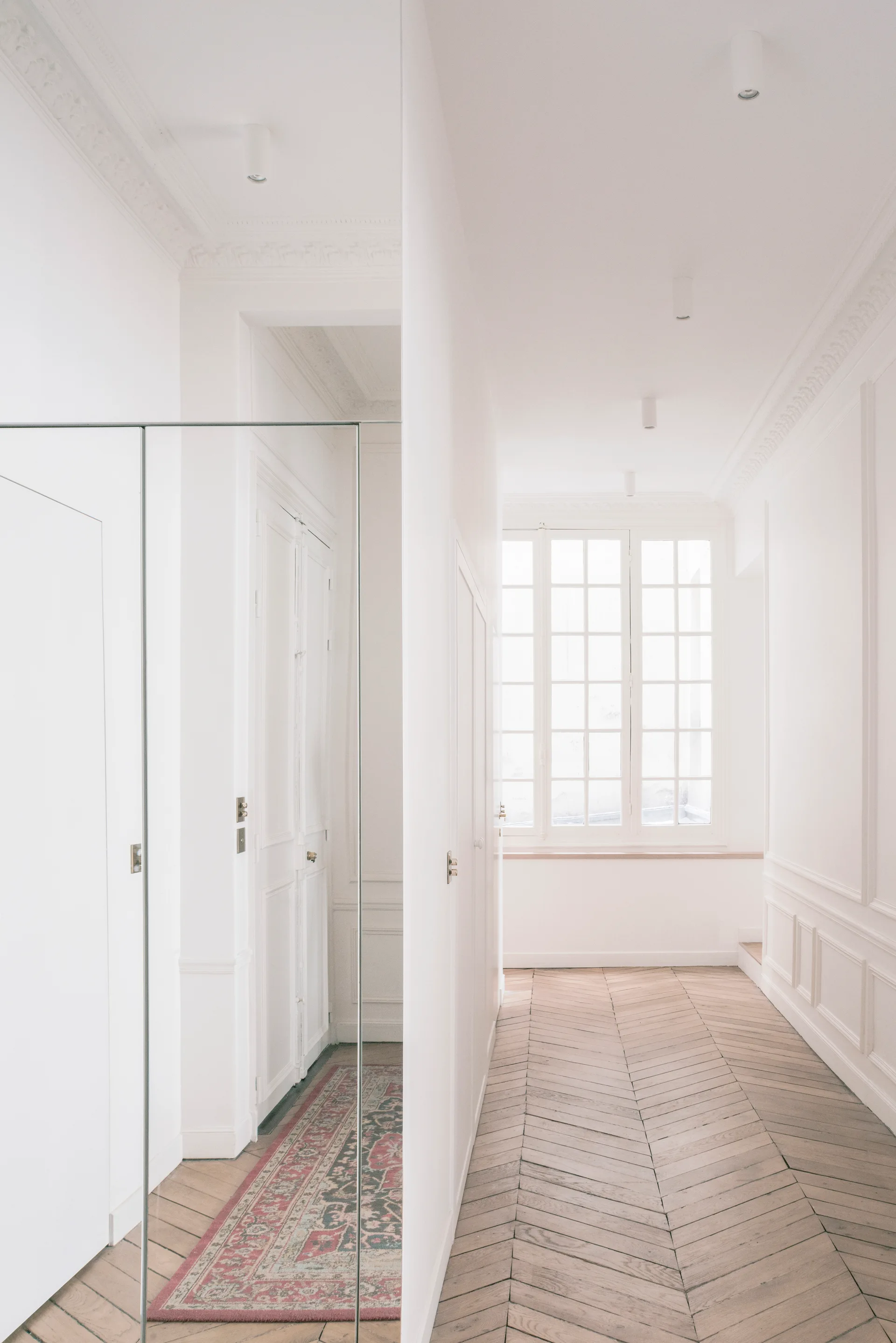 A bright white hallway with herringbone wood floors, classical wall molding, a tall multi-pane window, and a large mirror reflecting a red patterned rug.