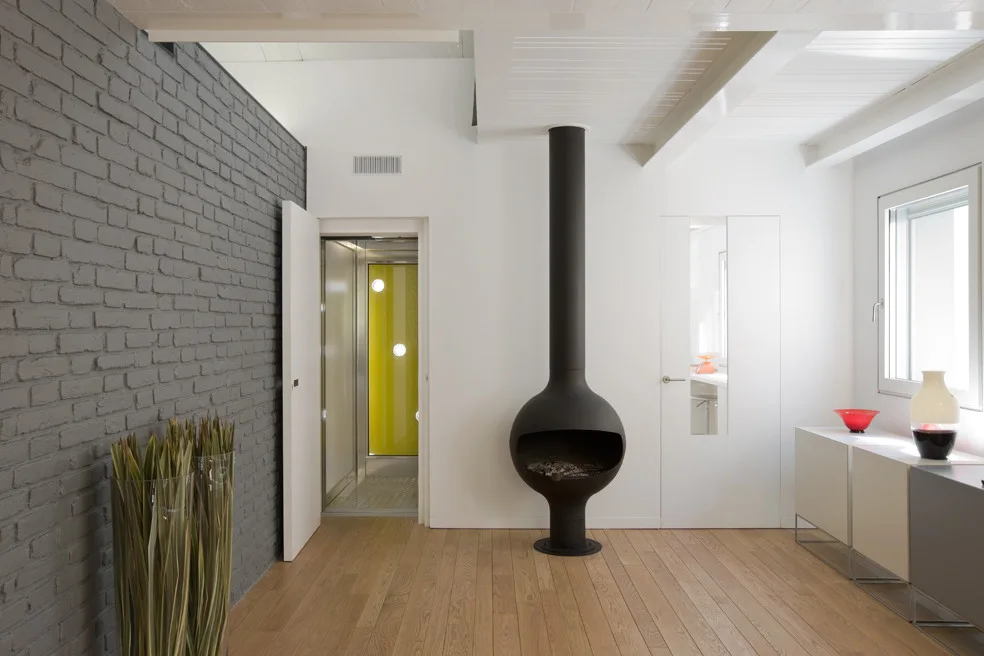 A modern living space features a sculptural black spherical fireplace, an exposed grey painted brick wall, and light wood flooring.