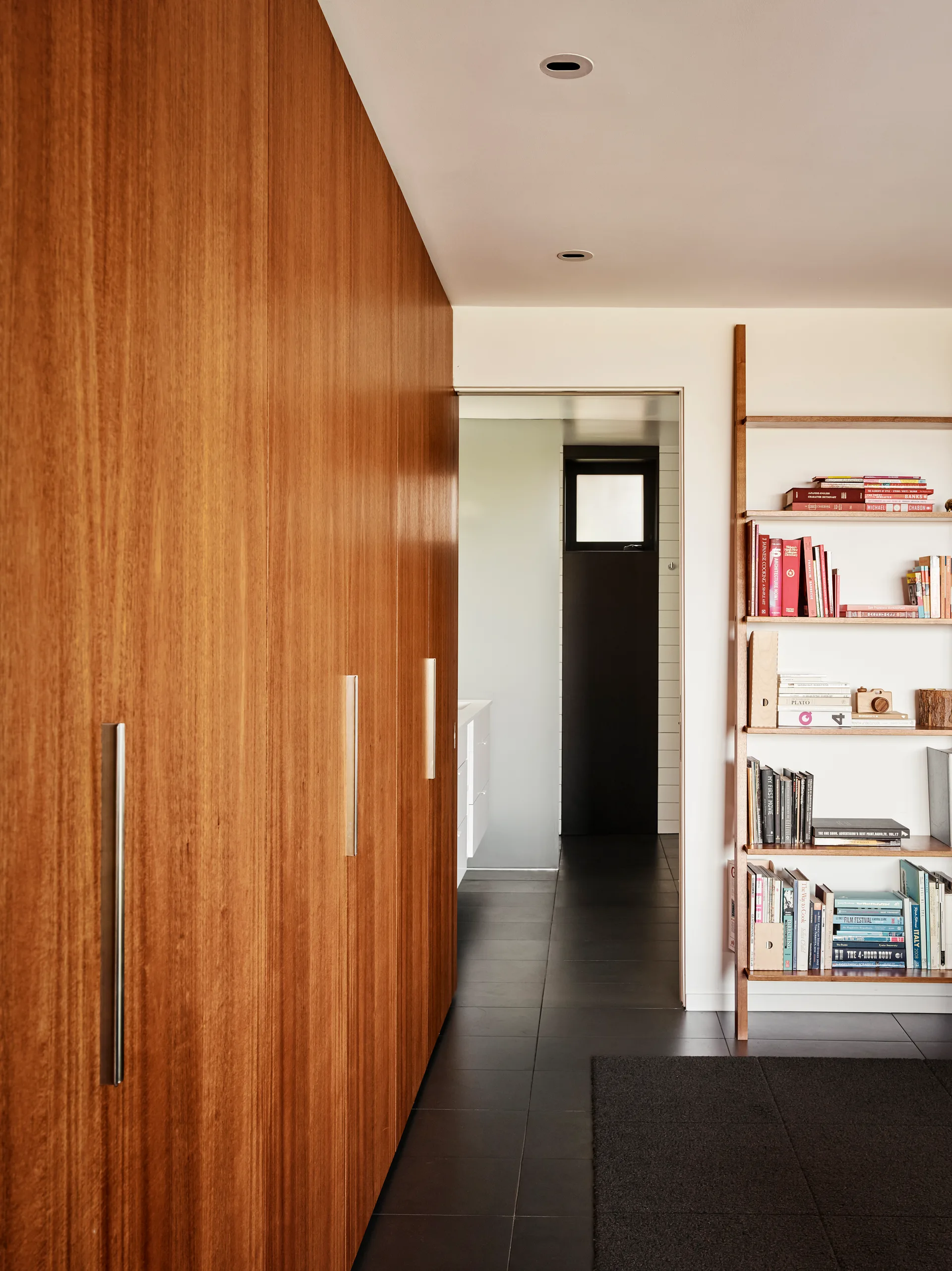 A modern hallway features a full wall of warm wood cabinets with silver handles, dark tiled floors, and a wooden ladder bookshelf filled with colorful books.