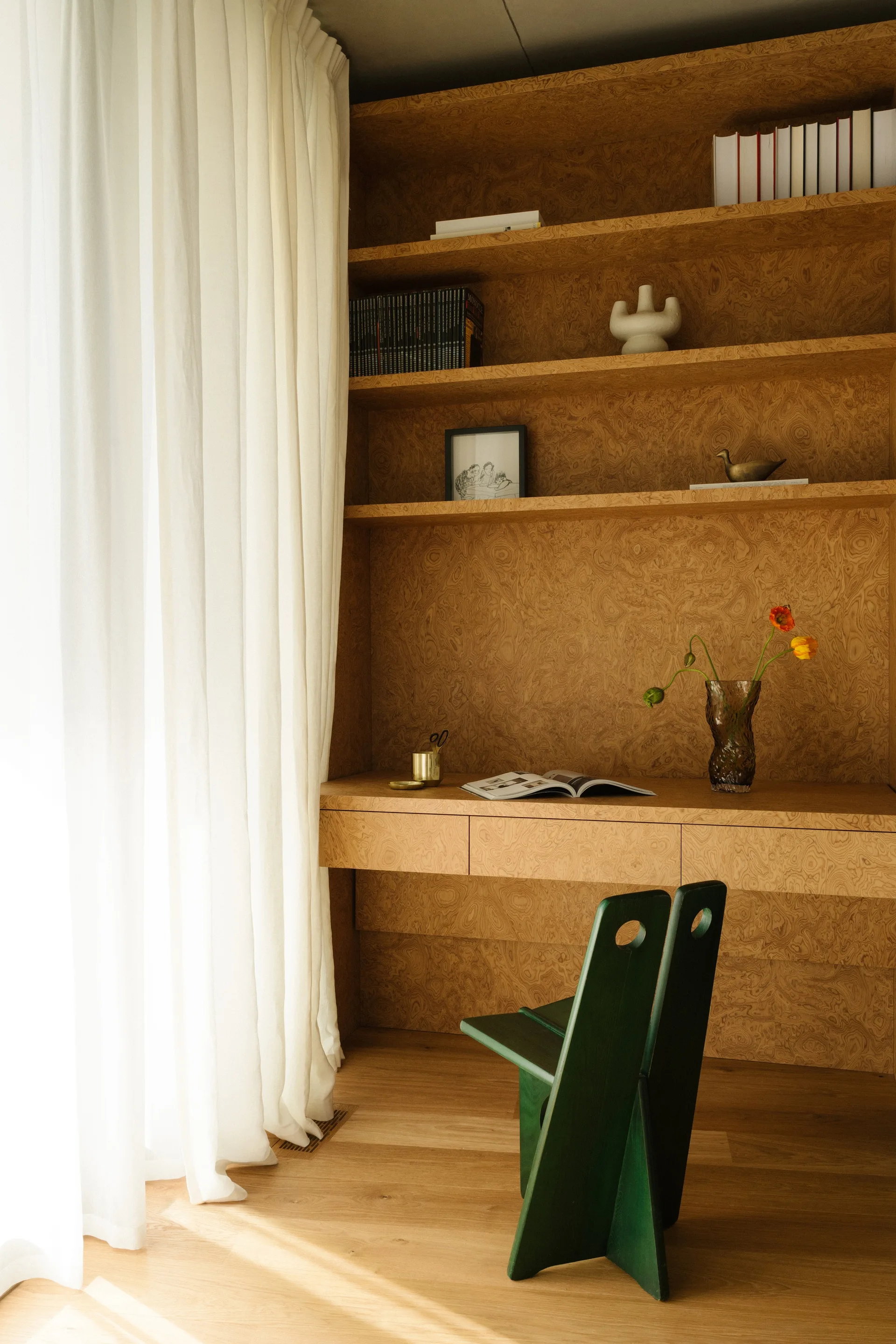 A home office nook features a built-in burl wood desk and shelves, a dark green sculptural chair, and white curtains framing natural light.