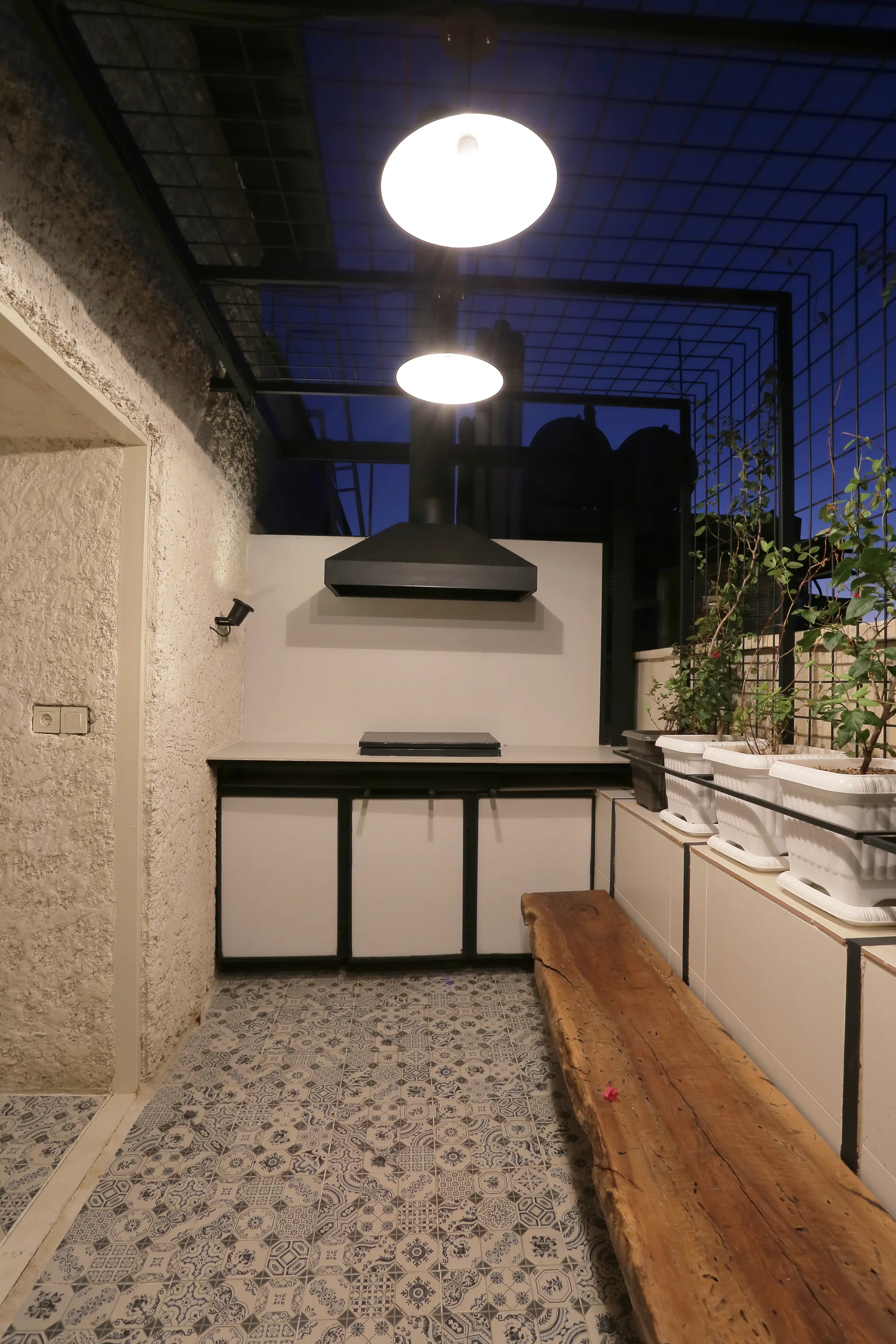 An outdoor kitchen featuring white cabinets, a black range hood, a rustic live-edge wooden bench, and patterned floor tiles under a metal grid ceiling.