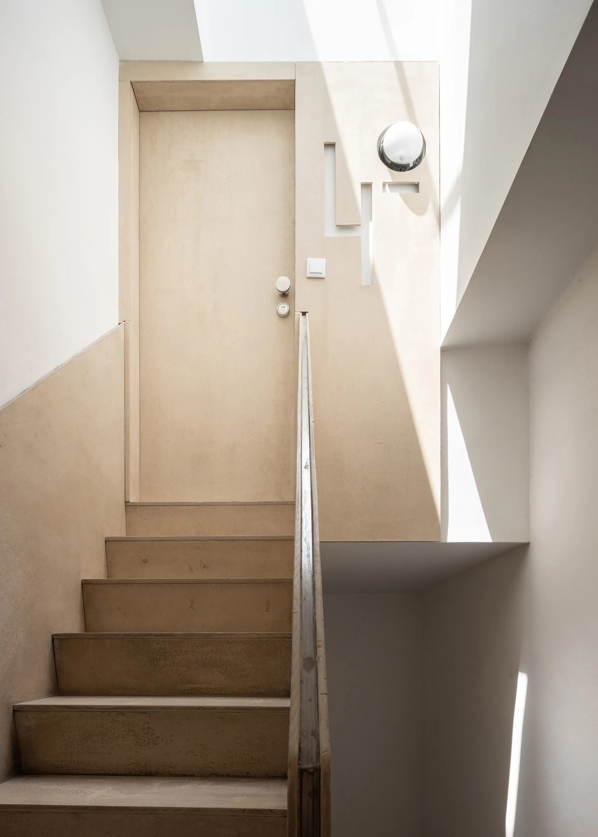 A minimalist wooden staircase leads to a matching wood door and wall panel illuminated by an overhead skylight.