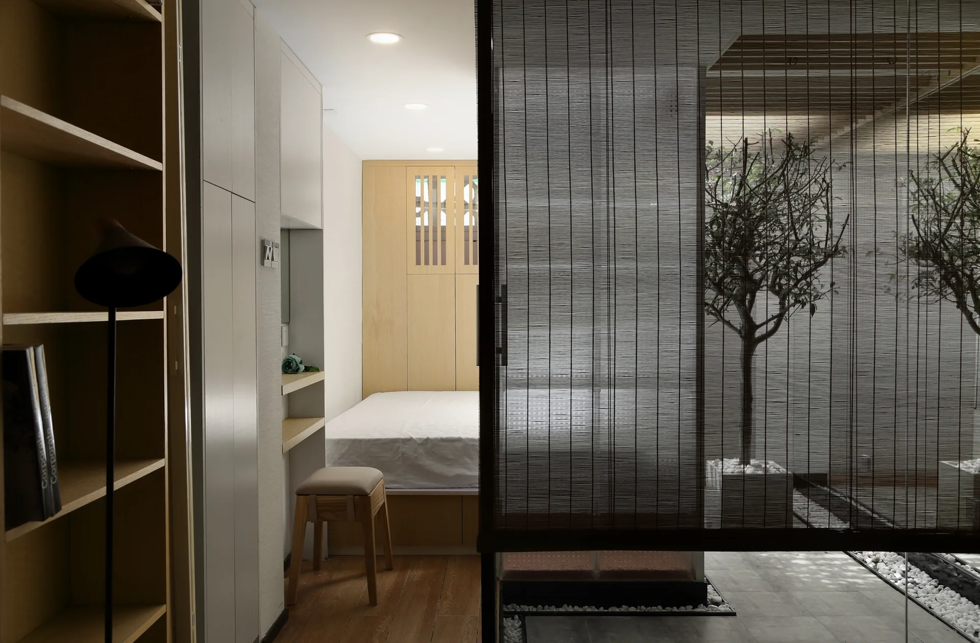 A minimalist bedroom featuring integrated light wood cabinets next to a dark slatted screen looking out onto a modern gravel courtyard.