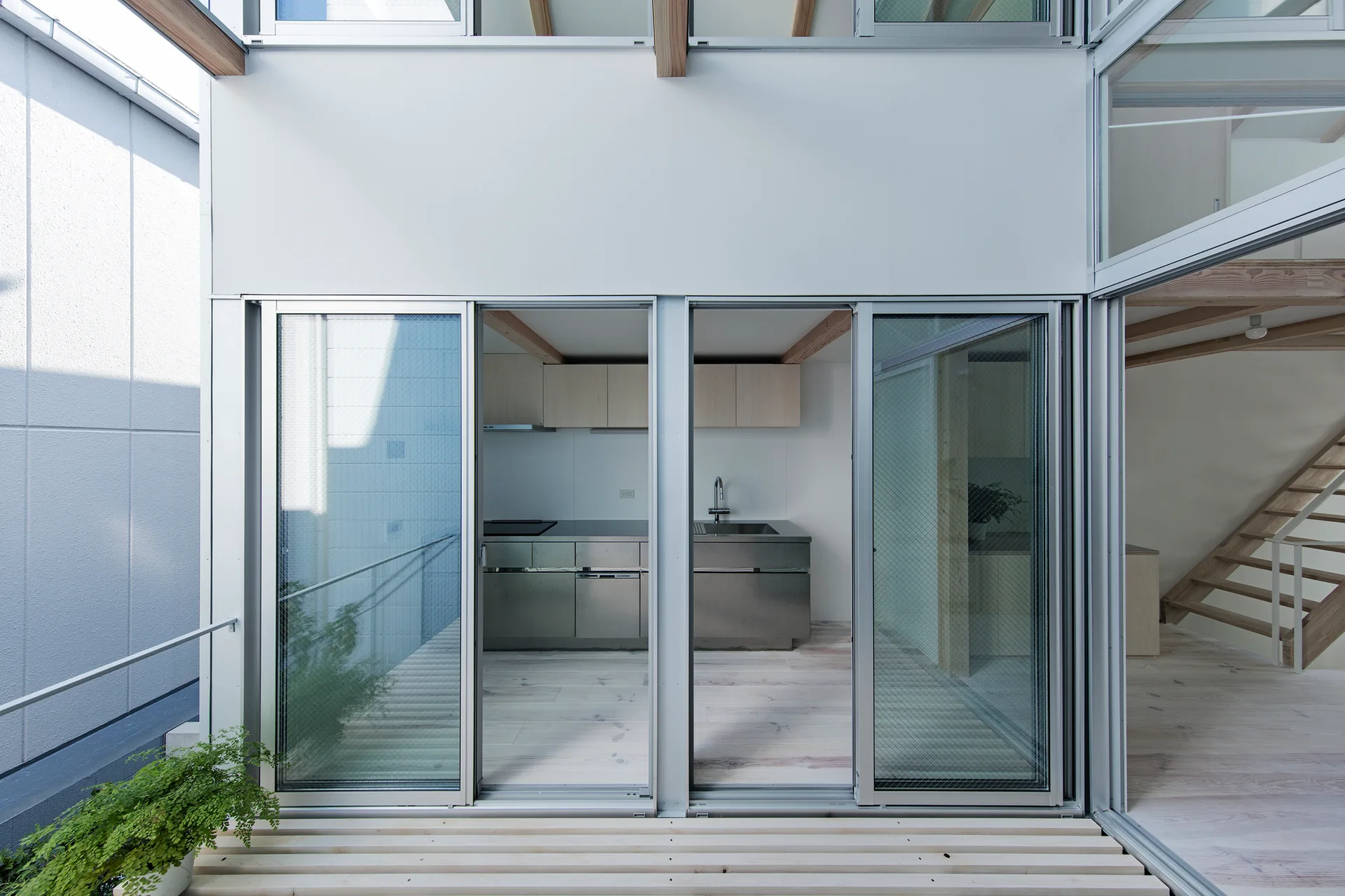 A minimalist kitchen with stainless steel lower cabinets and light wood upper cabinets is visible through glass sliding doors next to a wooden staircase.