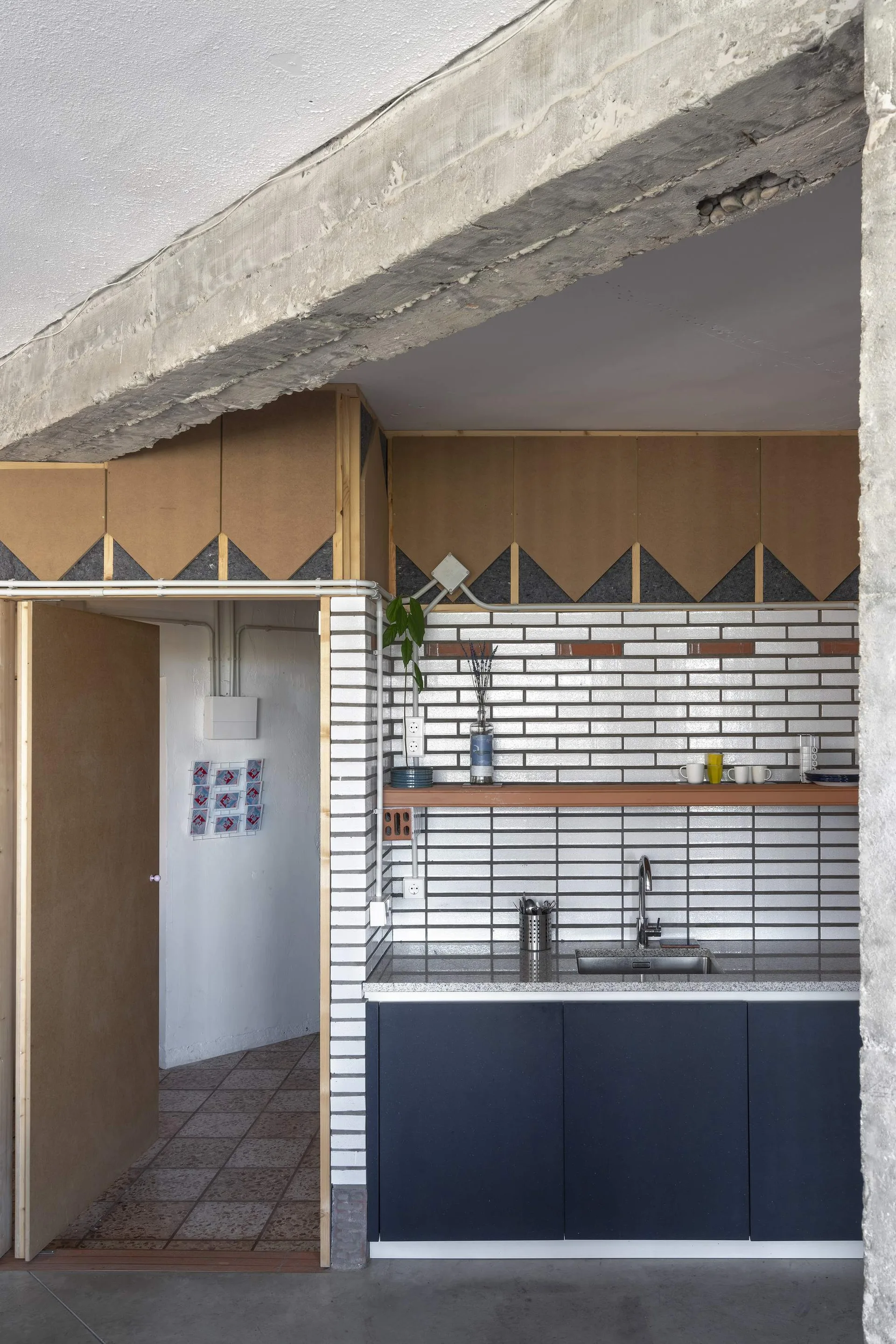 A kitchen with dark blue lower cabinets, a white tiled backsplash, a wooden floating shelf, and an exposed raw concrete ceiling beam.