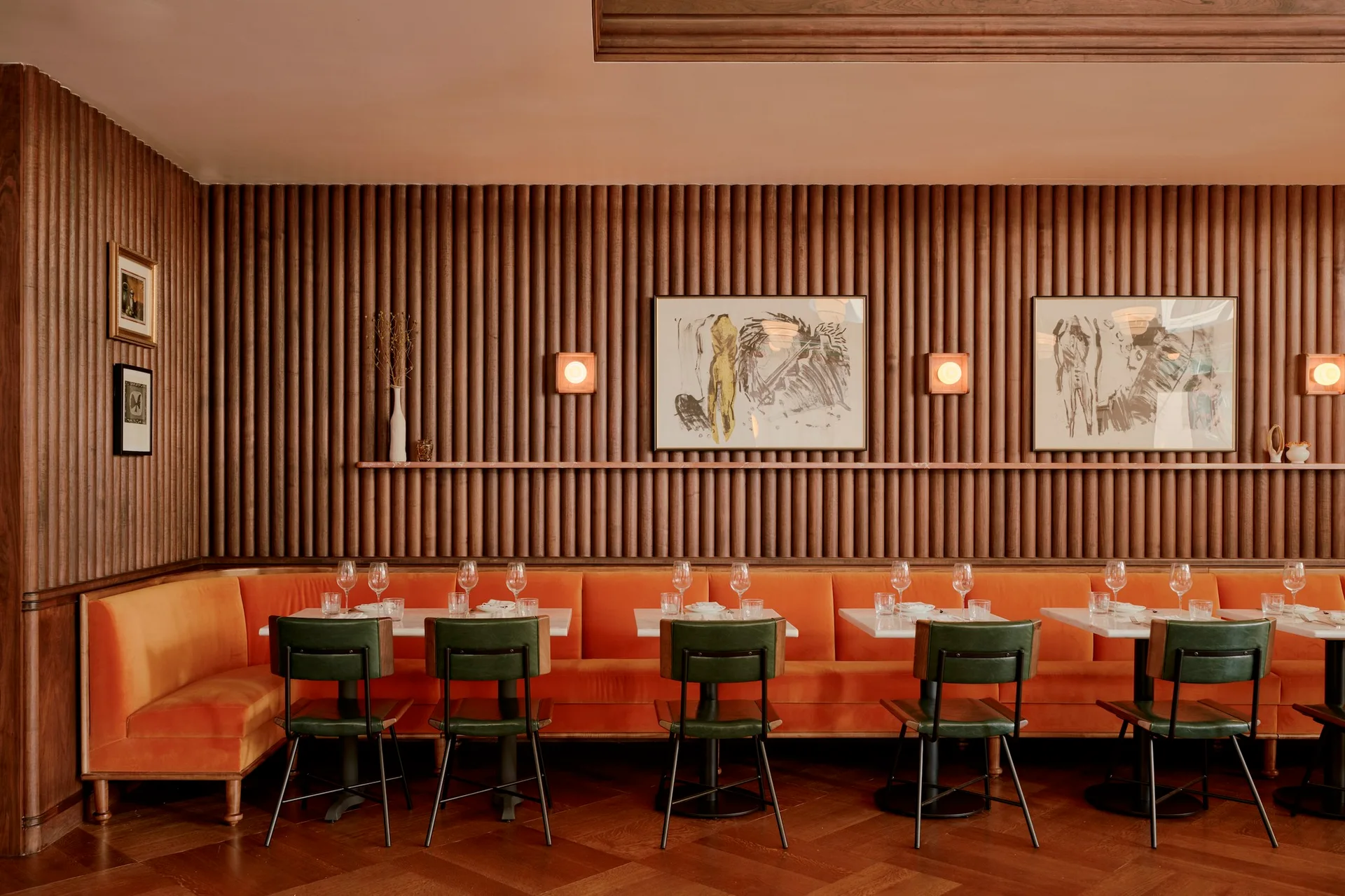 A modern dining area featuring a long orange velvet banquette, green upholstered chairs, and a vertical fluted wood accent wall.