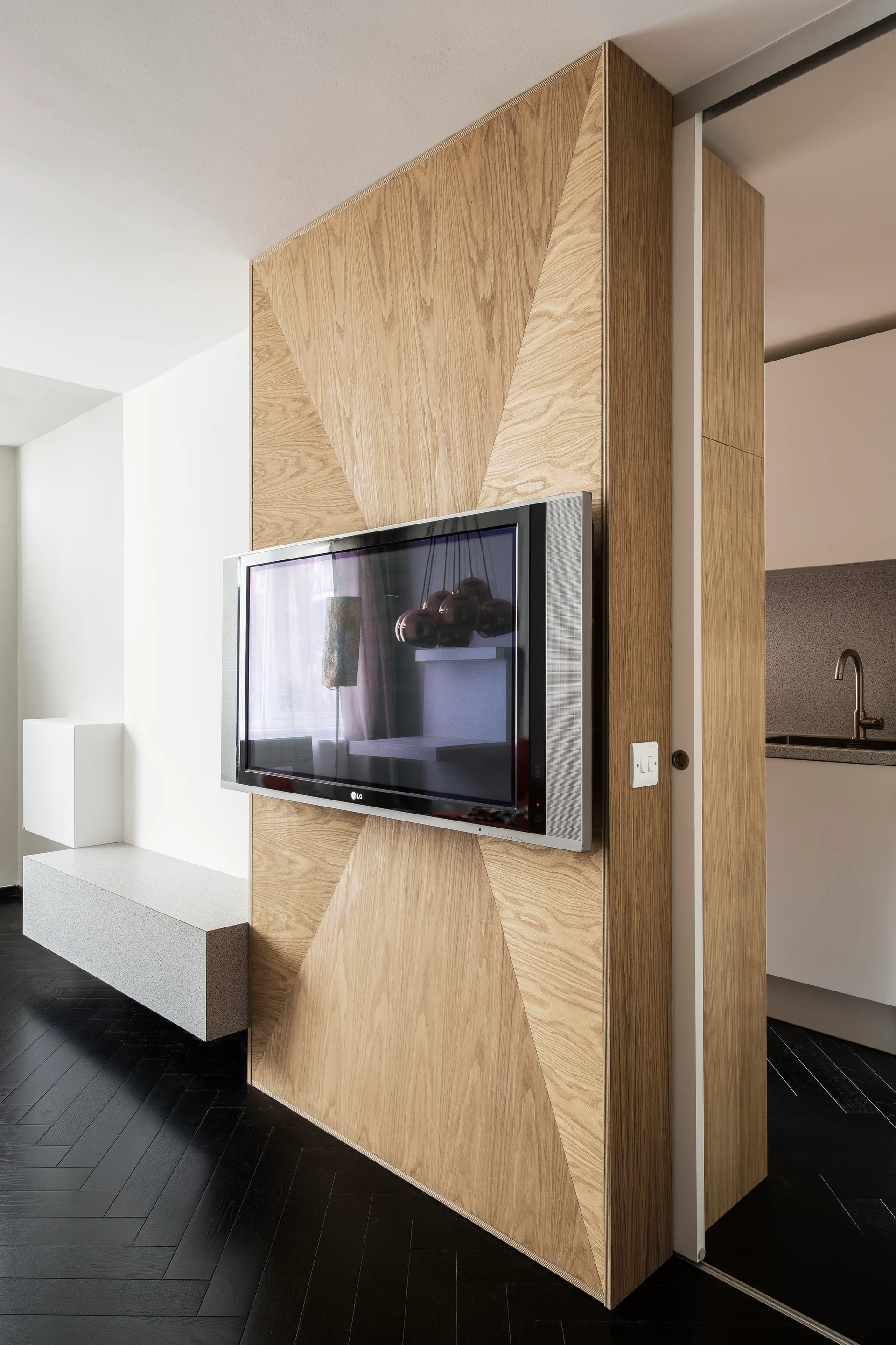 A wall-mounted television hangs on a custom geometric wood-paneled wall next to dark herringbone hardwood flooring and a floating grey stone bench.