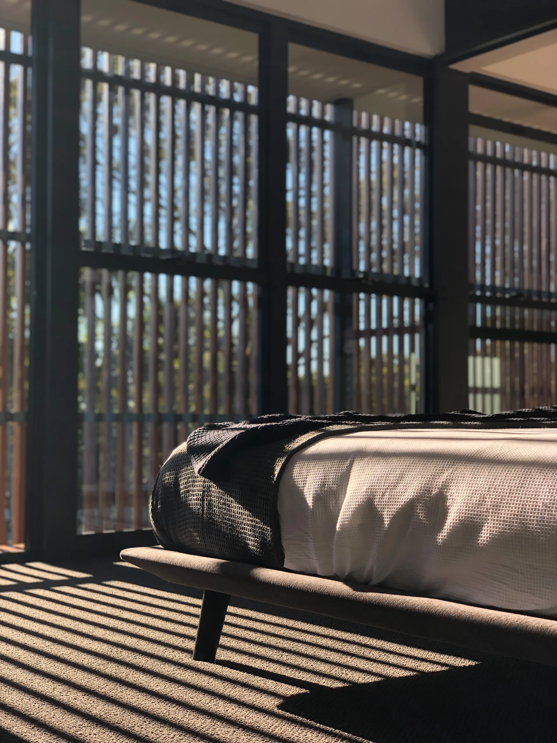 A low-profile bed with white and grey bedding sits next to floor-to-ceiling windows with vertical wood louvers casting striped shadows on the floor.