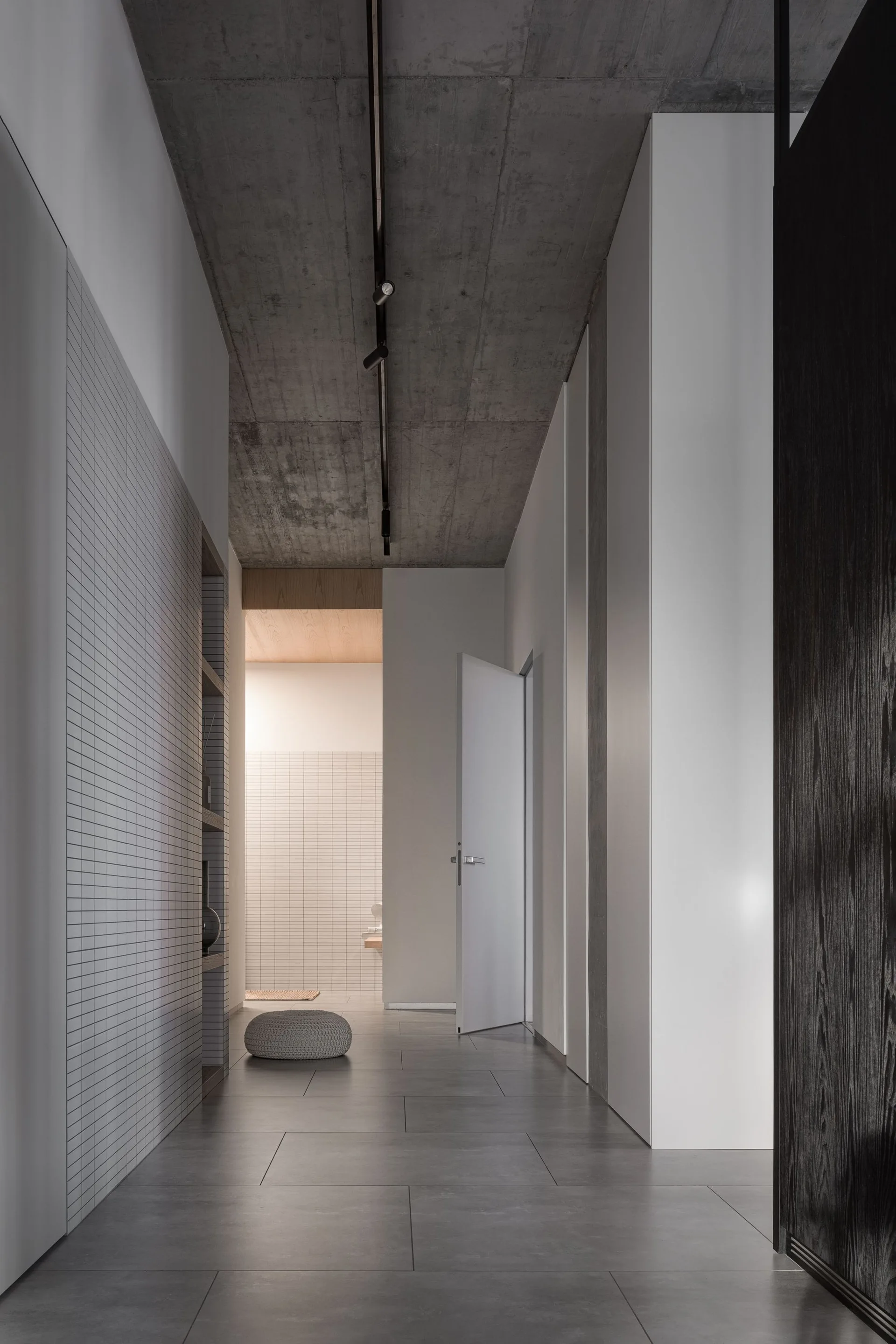 A long minimalist hallway features a raw concrete ceiling, a grid-patterned tile wall with recessed shelving, and large grey floor tiles.