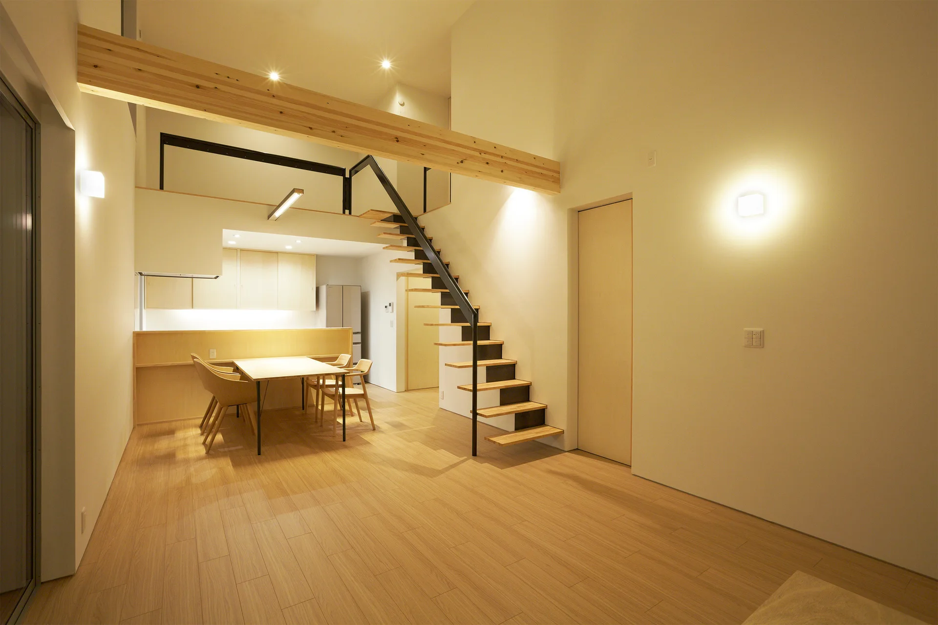 An open dining space features a light wood table set beneath a mezzanine loft with a black steel floating staircase and an exposed timber beam.