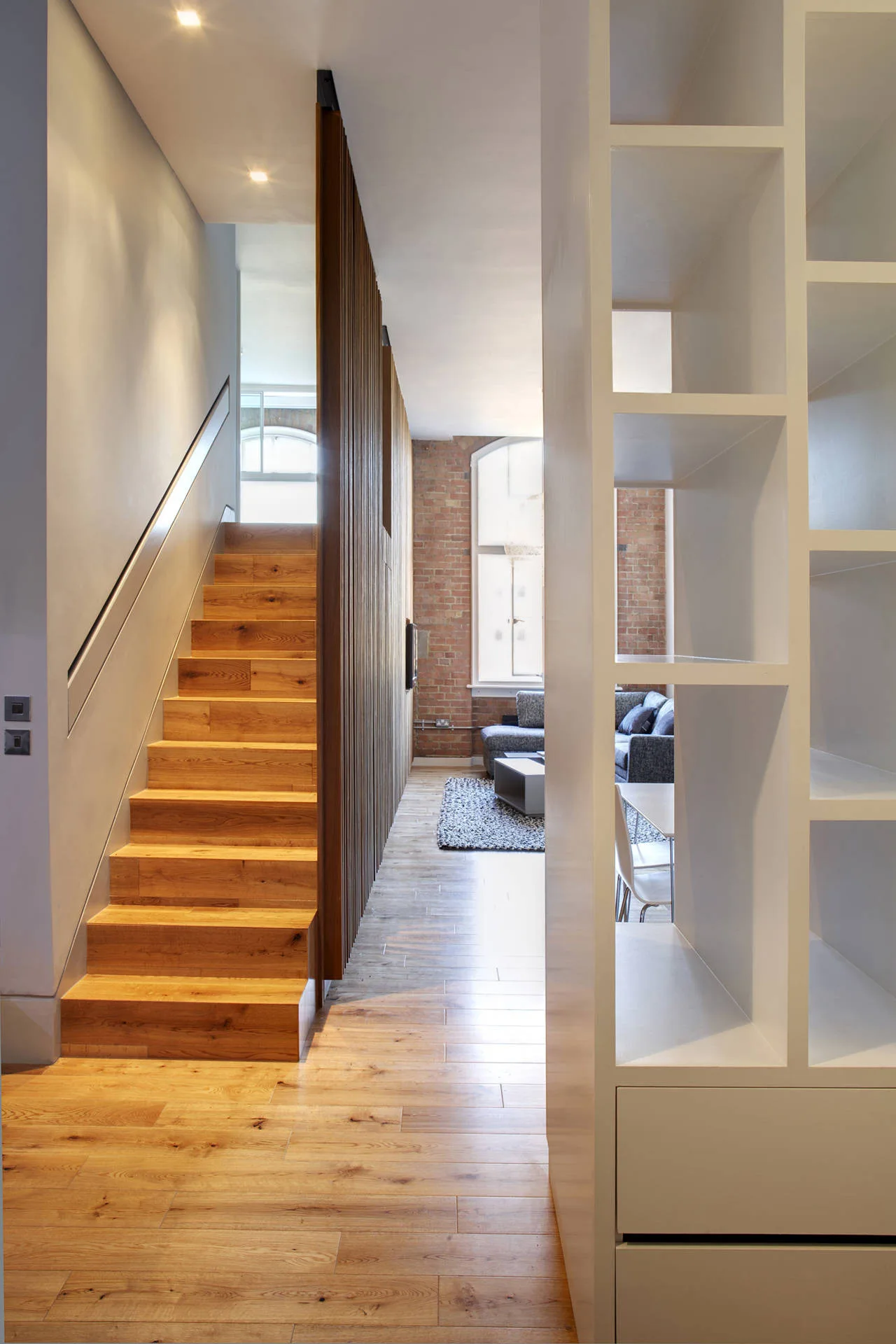 A natural wood staircase stands next to a vertical slatted partition leading into an open-plan loft living room with exposed brick walls.