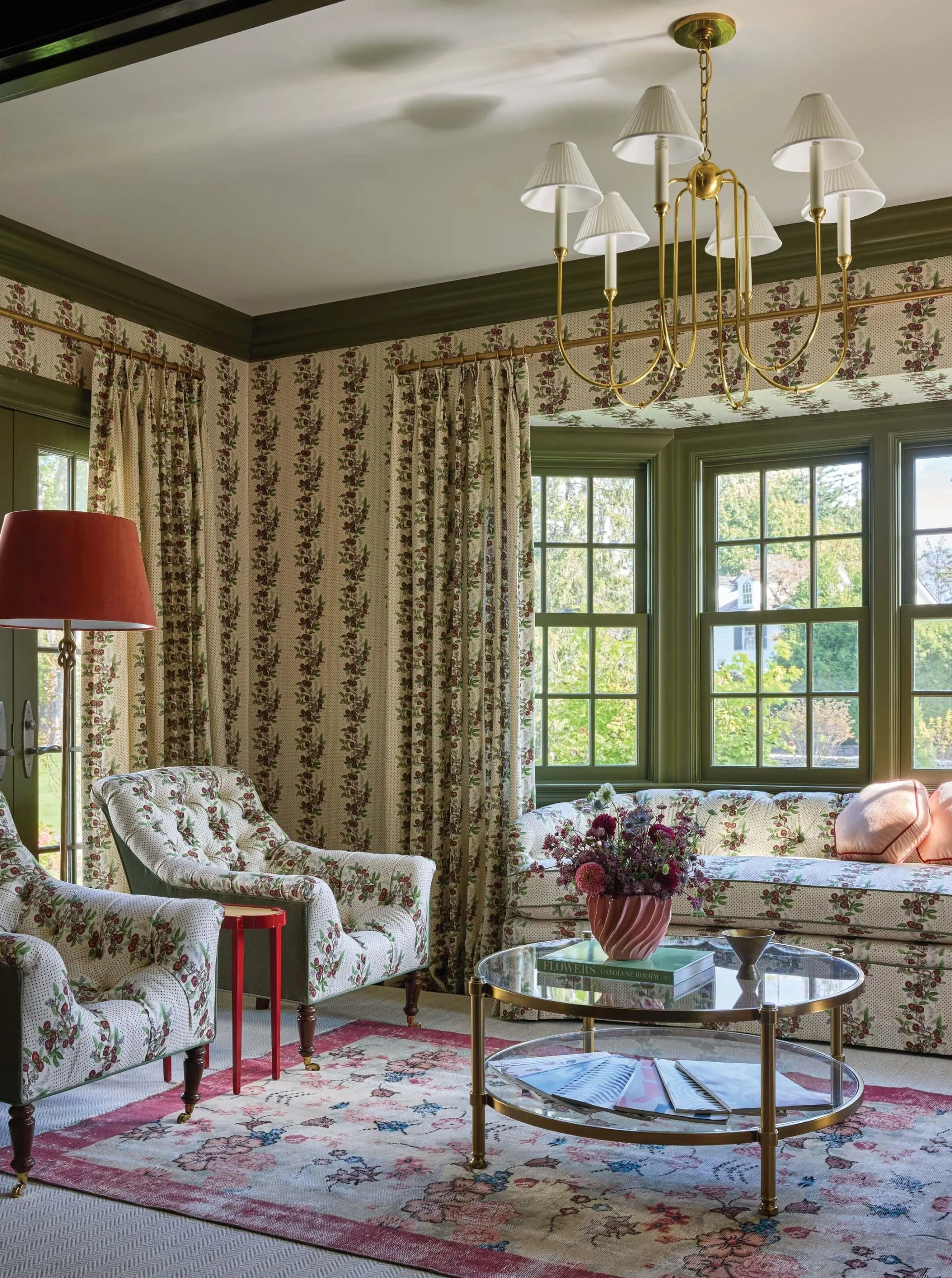 A traditional sitting room features matching floral wallpaper, drapery, and upholstered seating paired with olive green painted trim and a brass chandelier.