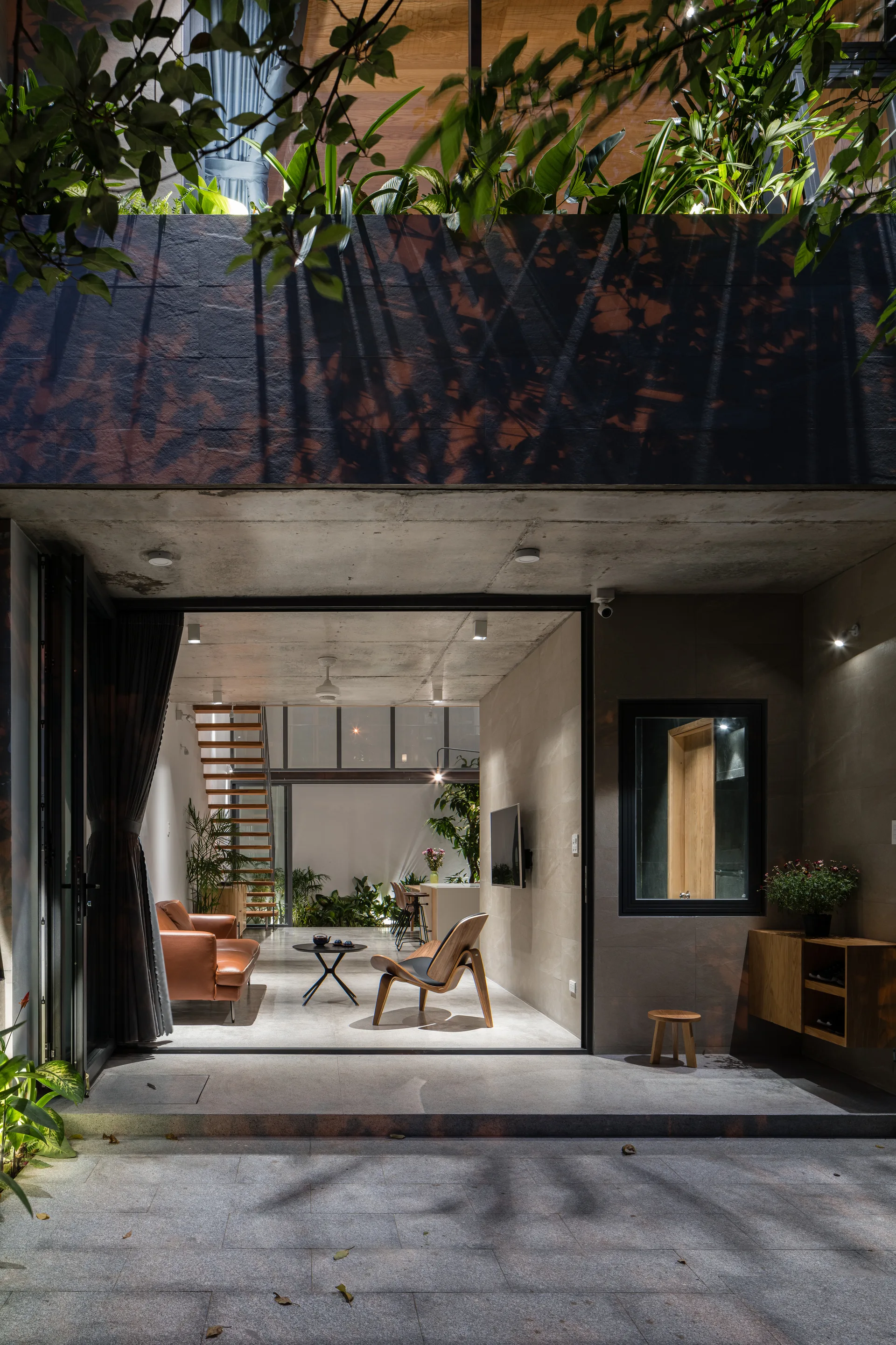 A modern open-plan living room with exposed concrete walls, a leather sofa, and a wooden staircase, opening directly onto a stone patio.