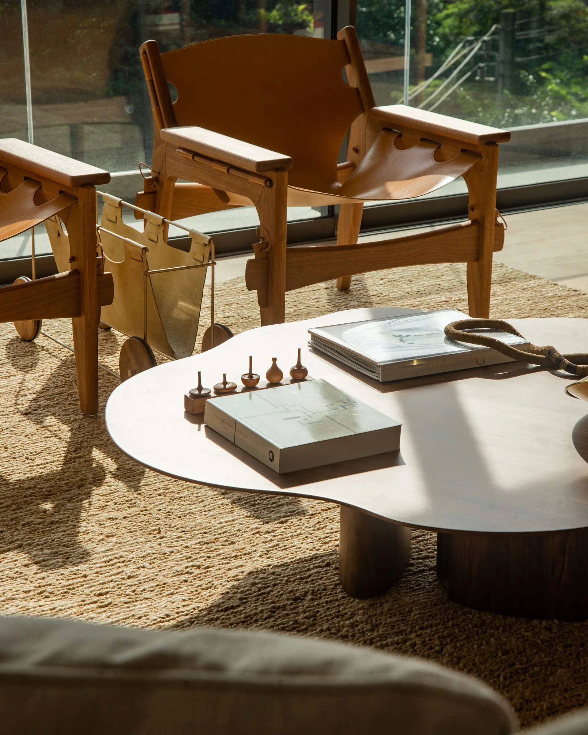 Two tan leather sling armchairs sit on a woven jute rug next to an organic wooden coffee table in a sunlit living room.