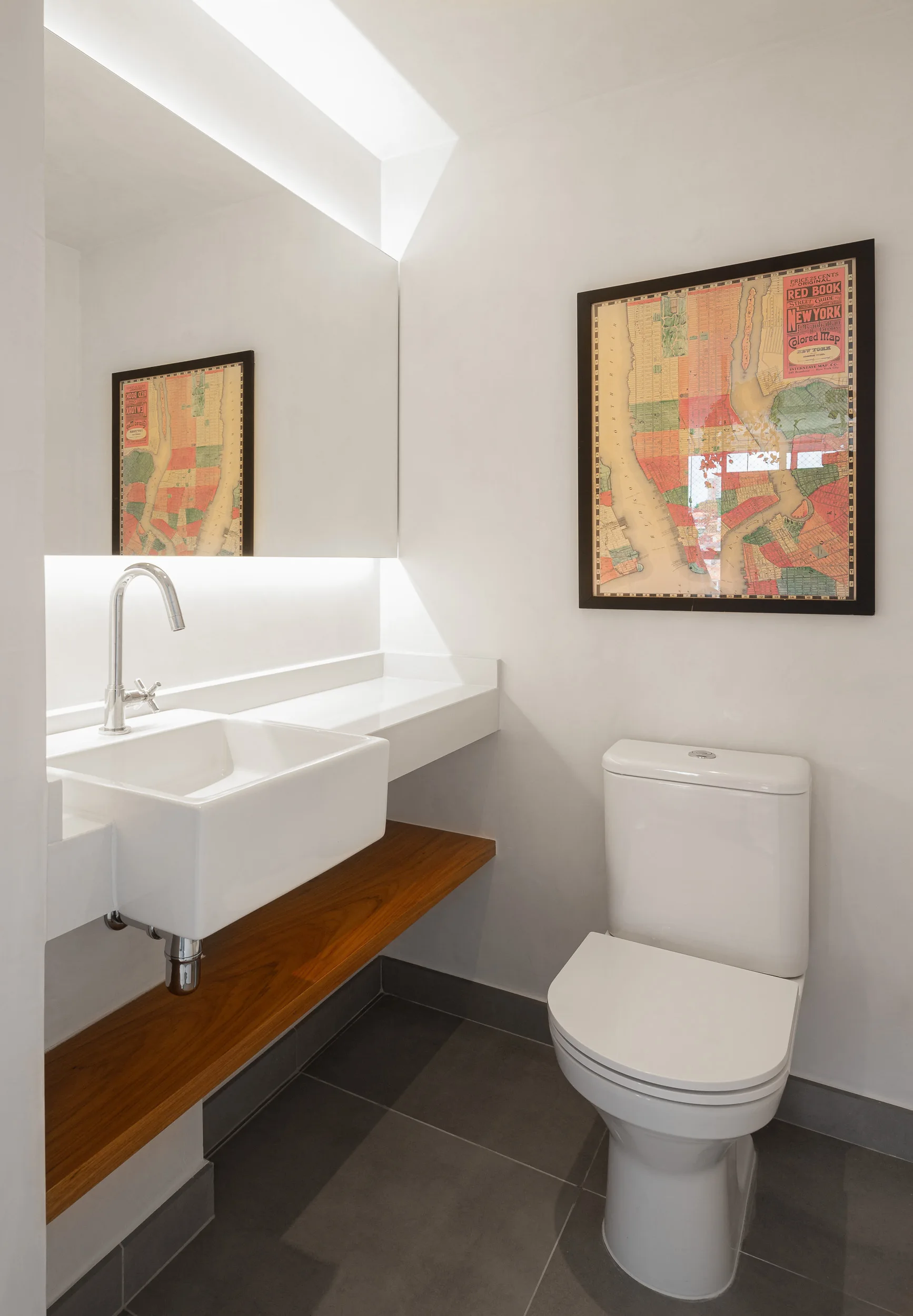 A minimalist powder room with a white toilet, a floating sink with a warm wood shelf underneath, and a framed vintage map on the wall.