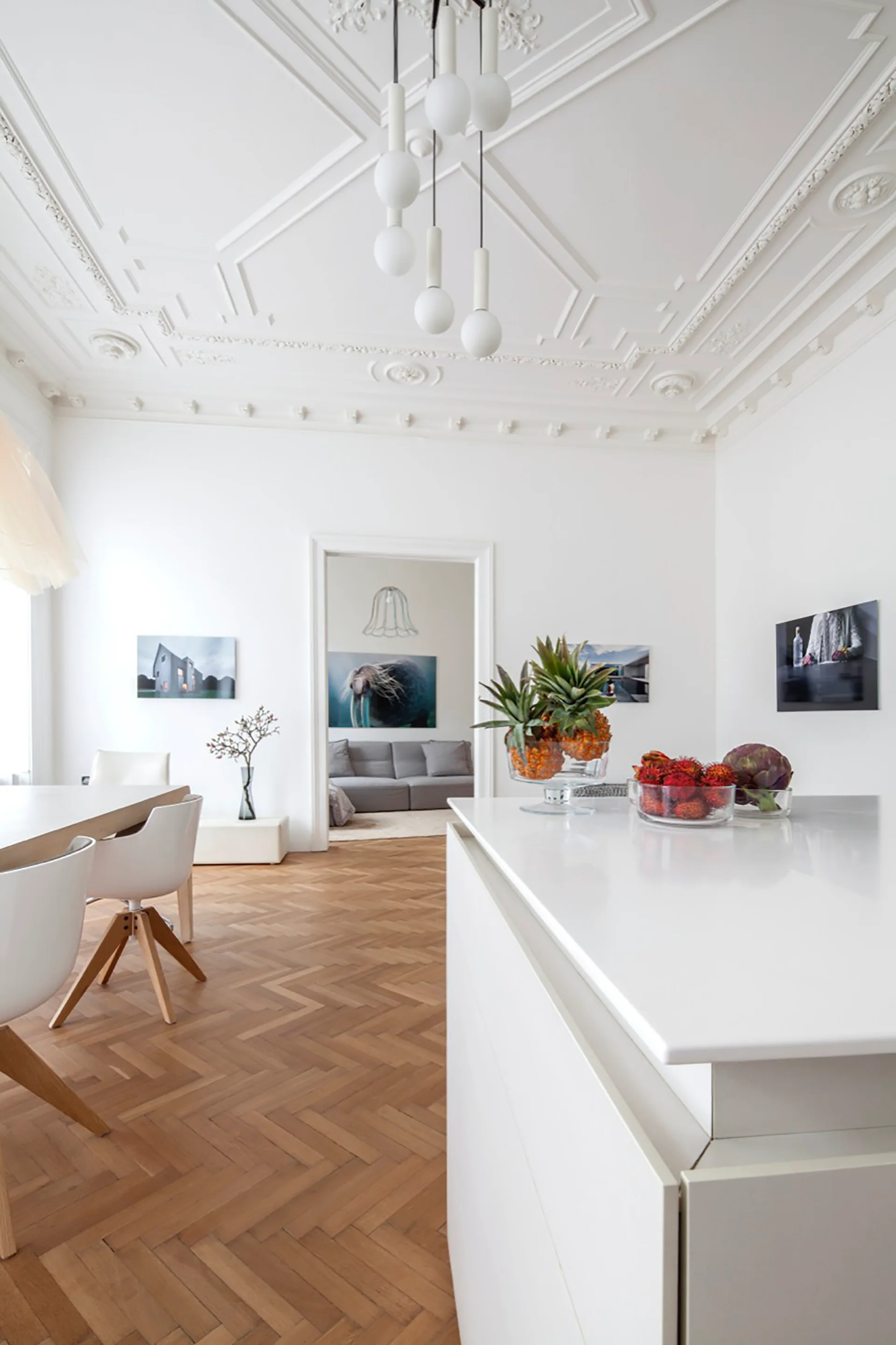 A white kitchen island sits on herringbone hardwood floors next to a light wood dining table below an ornate plaster ceiling.