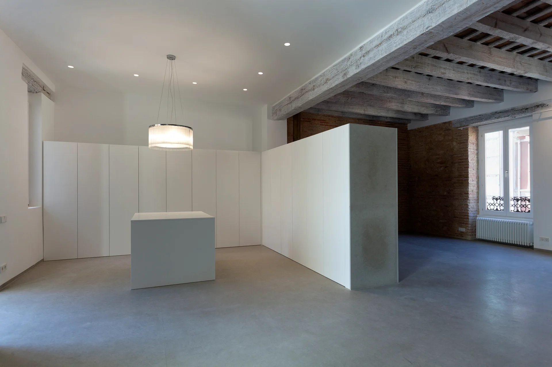 A minimalist room featuring a large wall of white modular cabinets, a central white island, exposed wood ceiling beams, and a concrete-look floor.