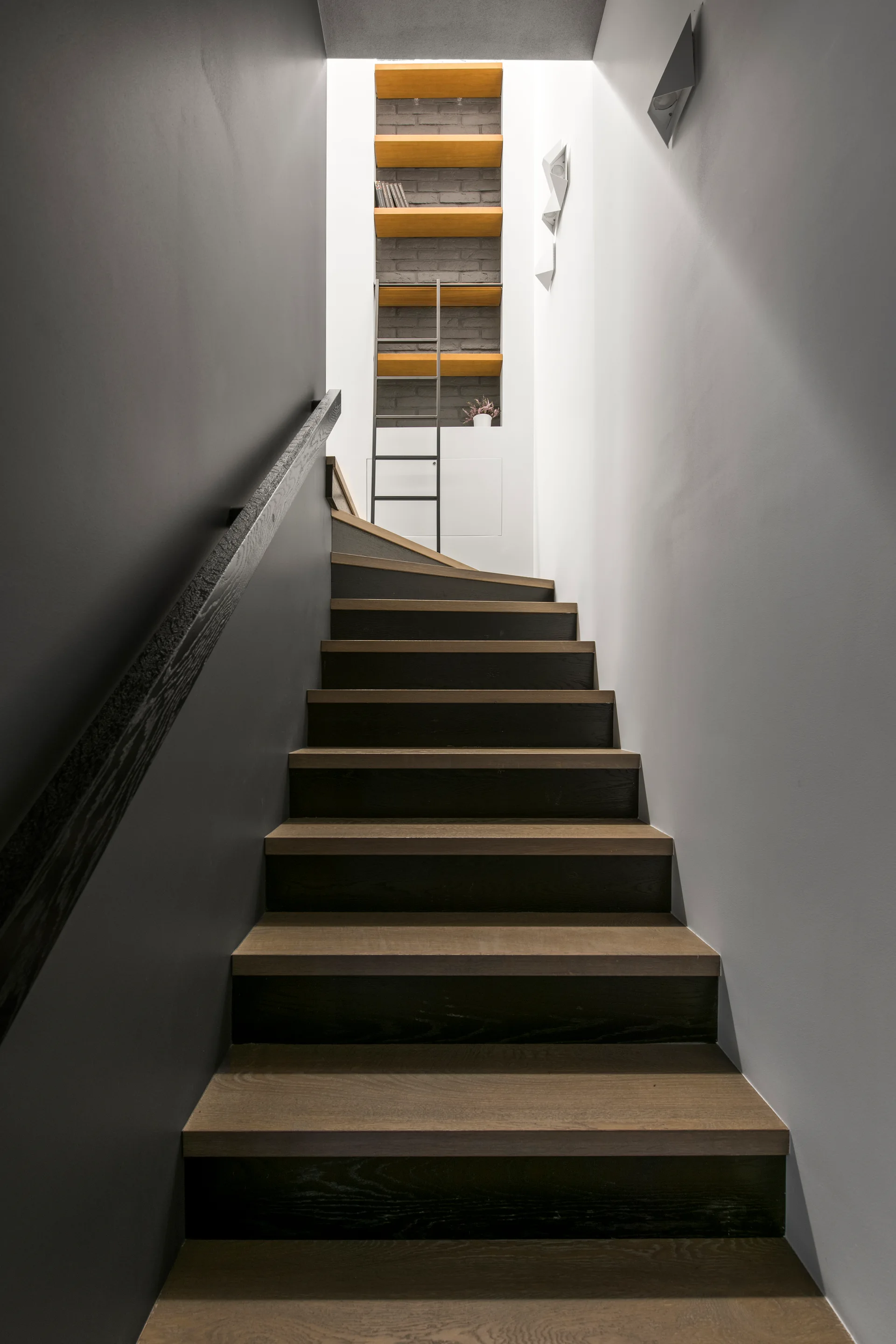 A modern wooden staircase ascends between gray walls toward a landing with a recessed vertical shelving unit and a metal ladder.