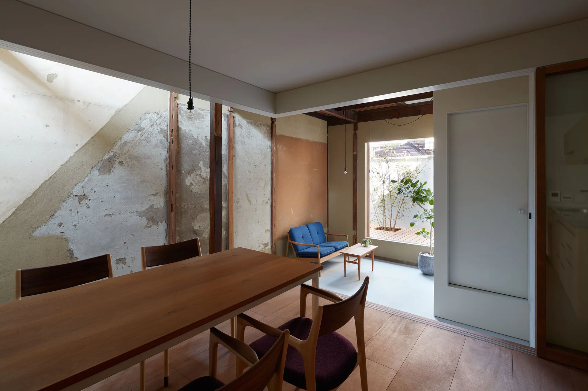 A minimalist dining area with a wooden table and chairs features an unfinished plaster wall beneath a skylight next to a small blue sofa.