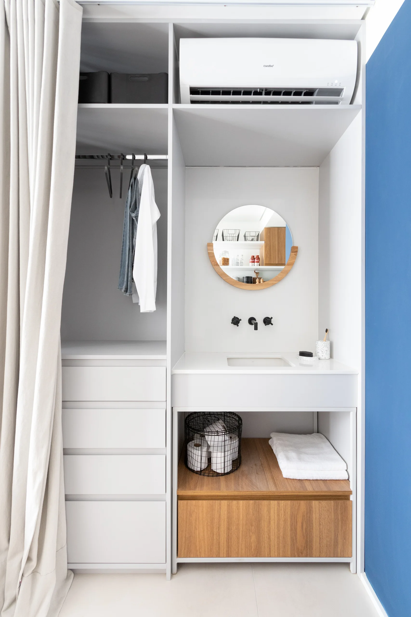 A white built-in unit combining a wardrobe closet with a bathroom vanity featuring a round mirror, black faucet, and wood accents.