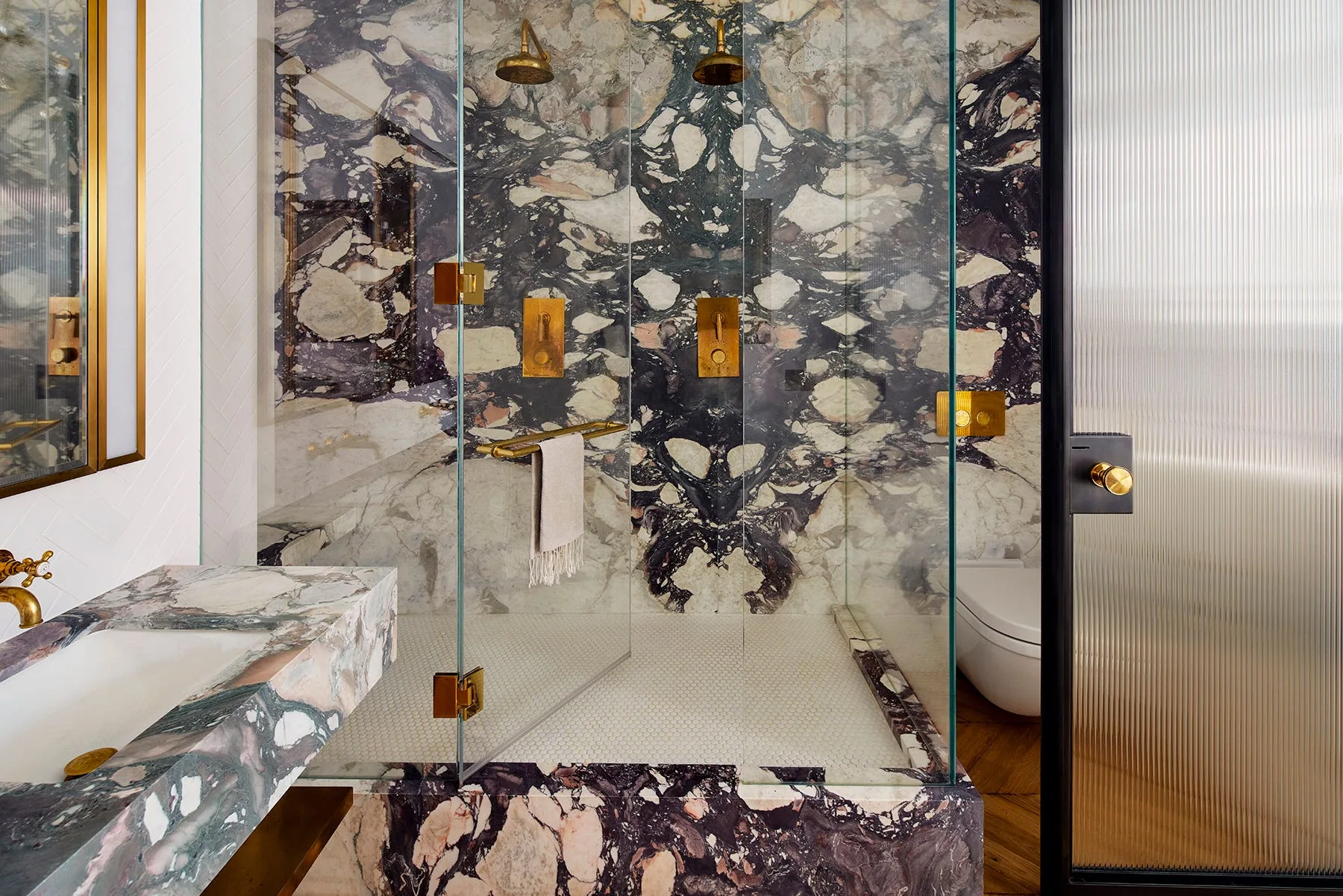 A glass enclosed walk-in shower with high-contrast book-matched purple marble walls and double brass shower heads.