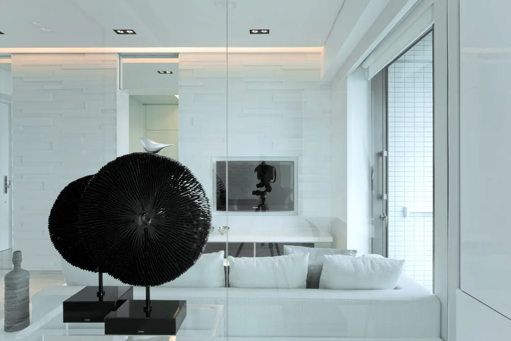 A minimalist white living room features a low white sofa, textured wall paneling, and two large black circular sculptures on a clear console table.