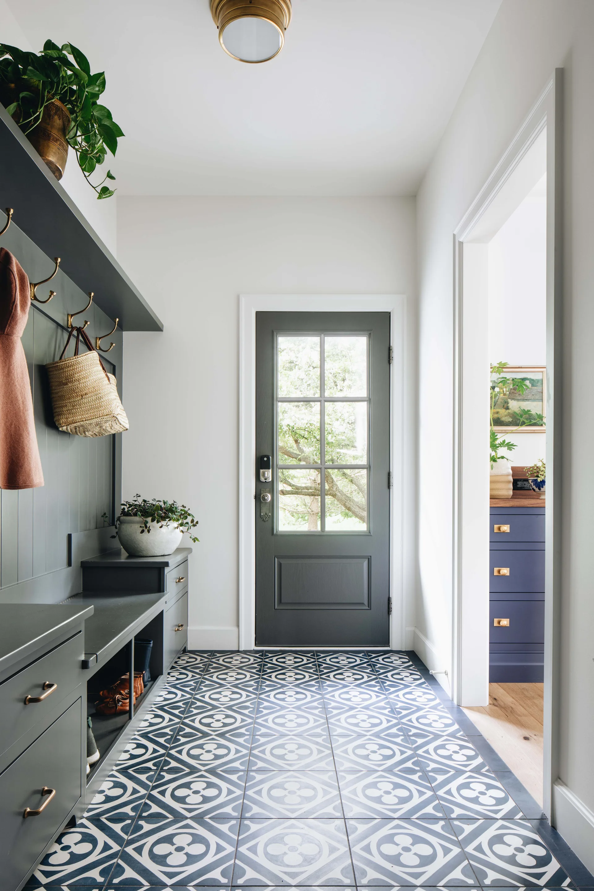 A bright mudroom with a dark green built-in storage bench, brass coat hooks, patterned floor tiles, and a gray entry door with glass panels.