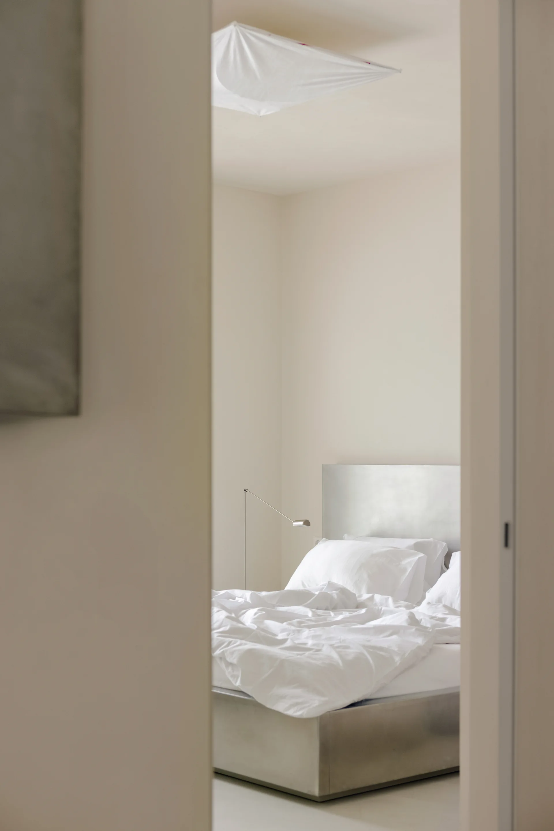 A minimalist bedroom seen through a doorway, featuring a bed with a brushed metal headboard, white linens, and a slim floor lamp.