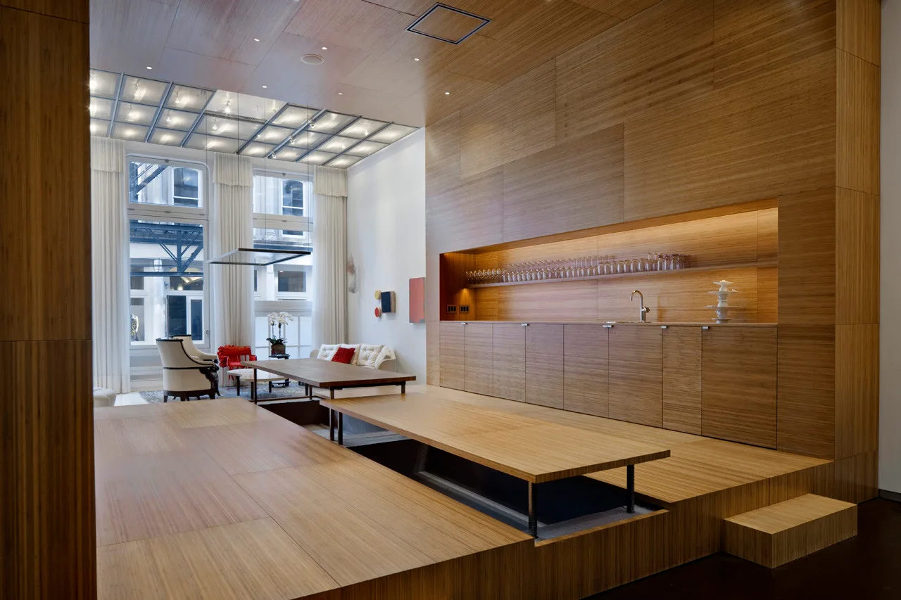A spacious modern loft featuring extensive light wood paneling on the walls, ceiling, and raised platforms, with a low dining table and recessed kitchen niche.