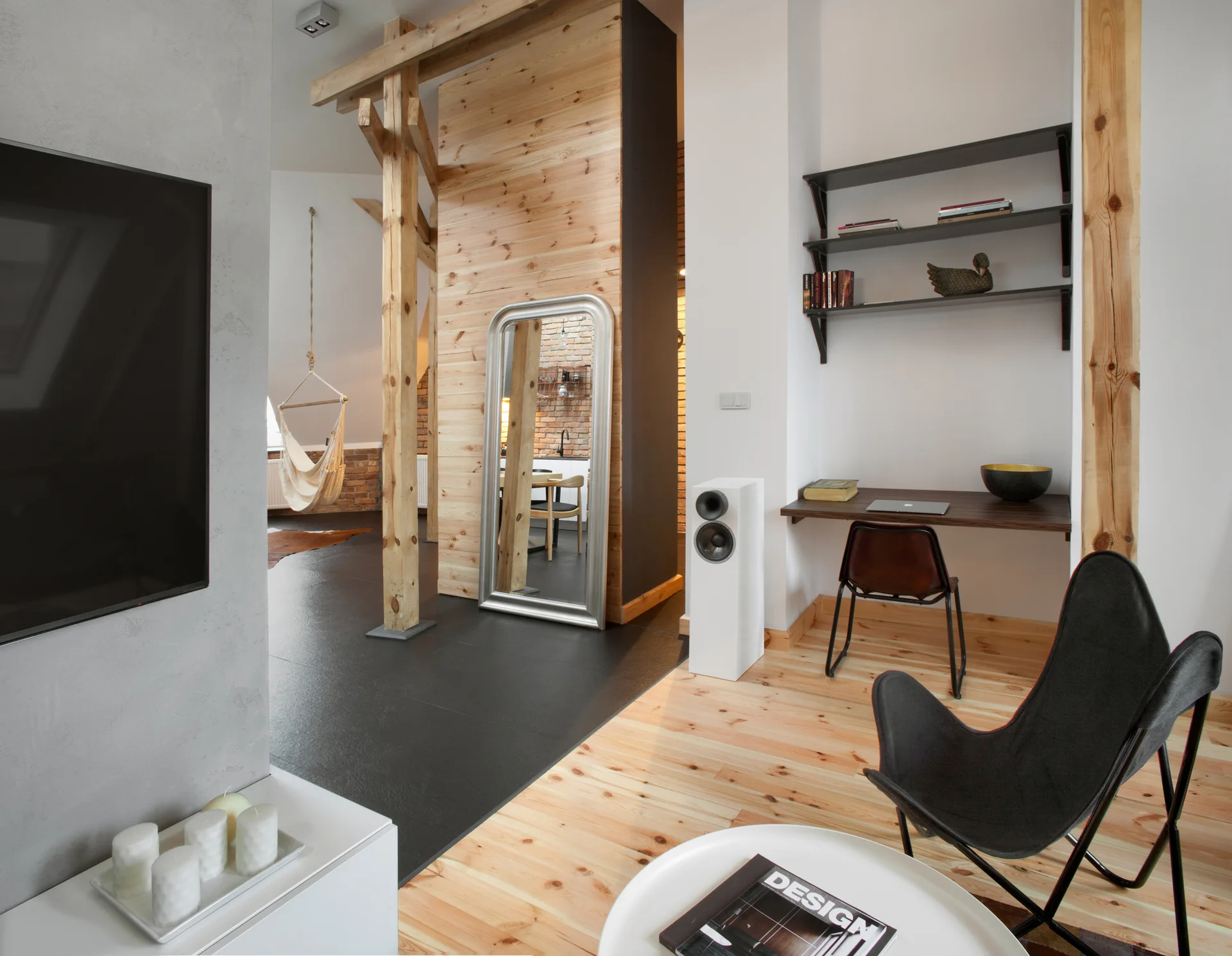 A modern loft interior featuring a wooden structural beam, a floating desk with wall shelves, a black butterfly chair, and mixed flooring transition.
