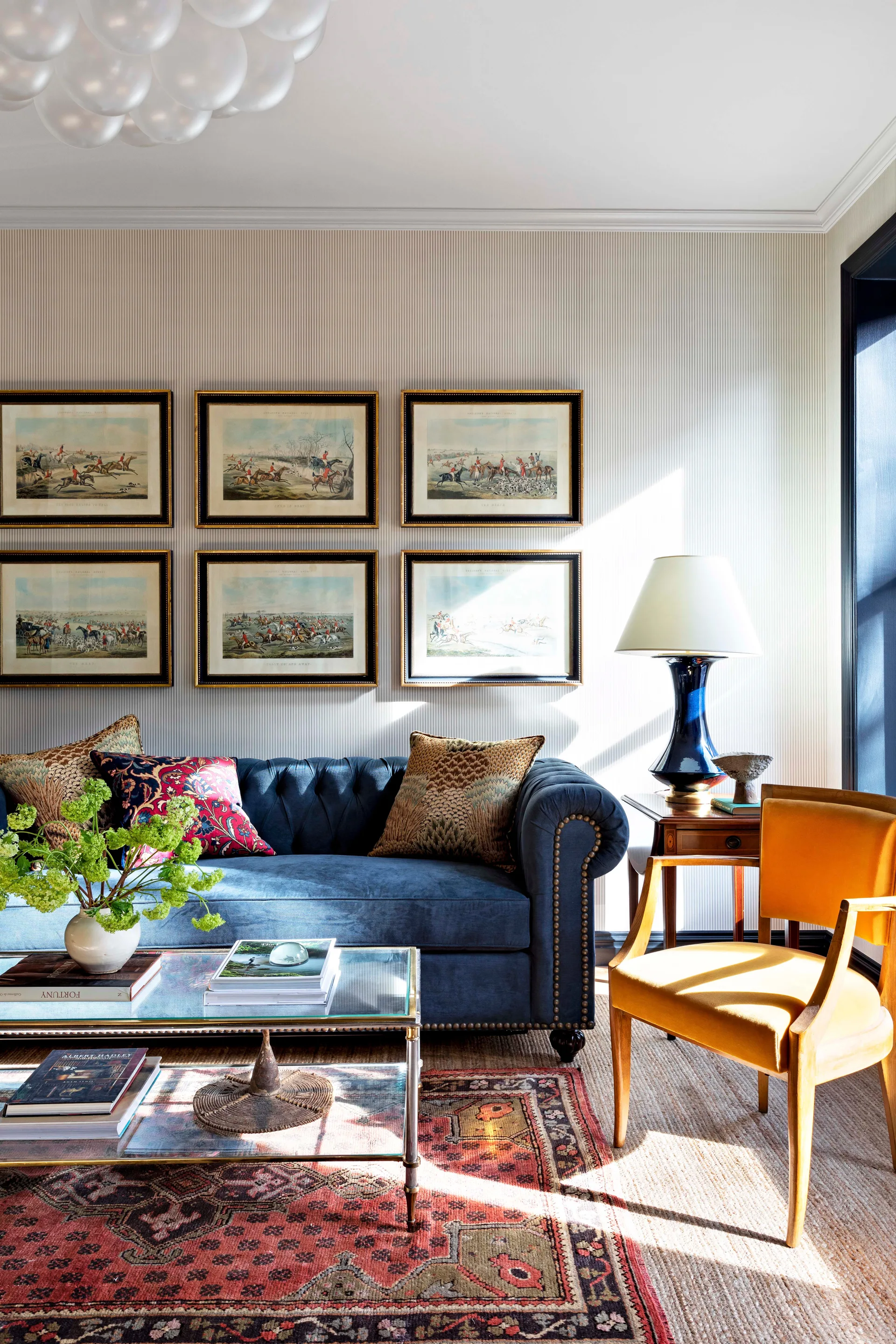A navy velvet tufted sofa is flanked by a mustard yellow armchair in a living room with pinstriped wallpaper and a grid of six framed prints.