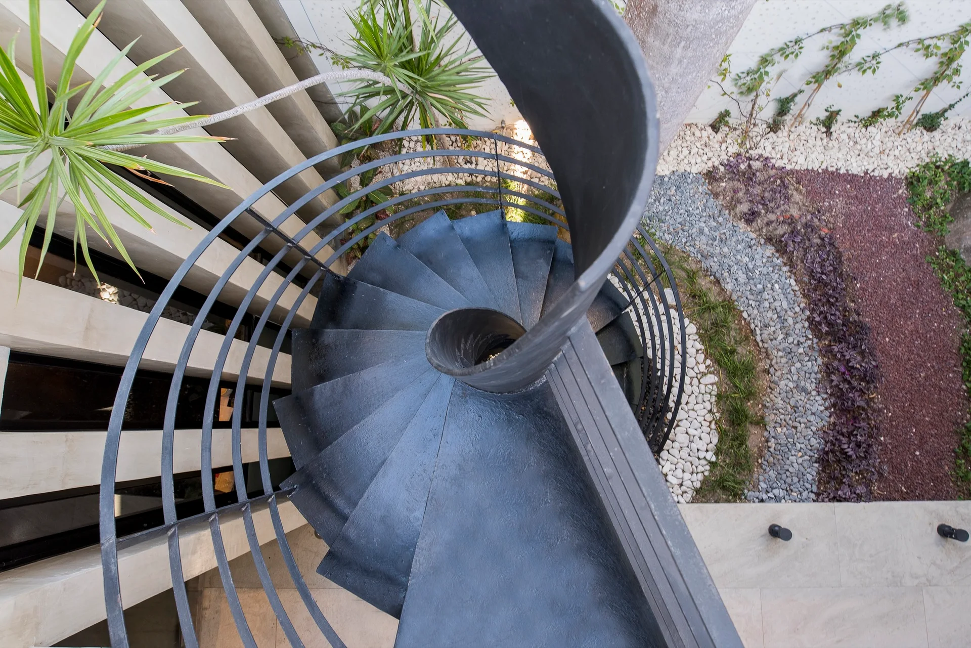 A top-down view of a black metal spiral staircase next to a beige slatted wall and a landscaped pebble garden.