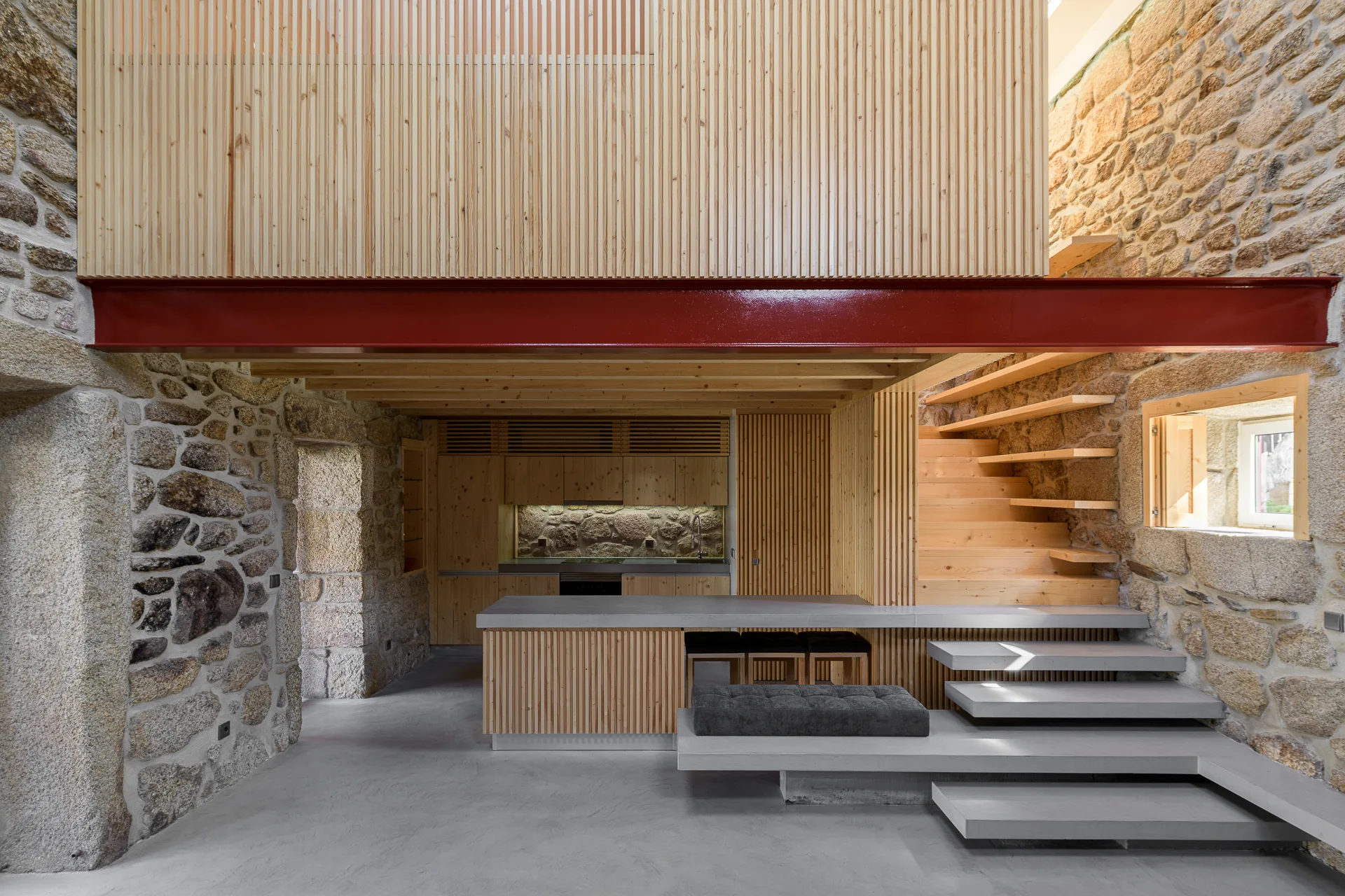 A modern rustic kitchen featuring rough exposed stone walls, vertical wood slat paneling, concrete platform steps, and a bright red steel beam.