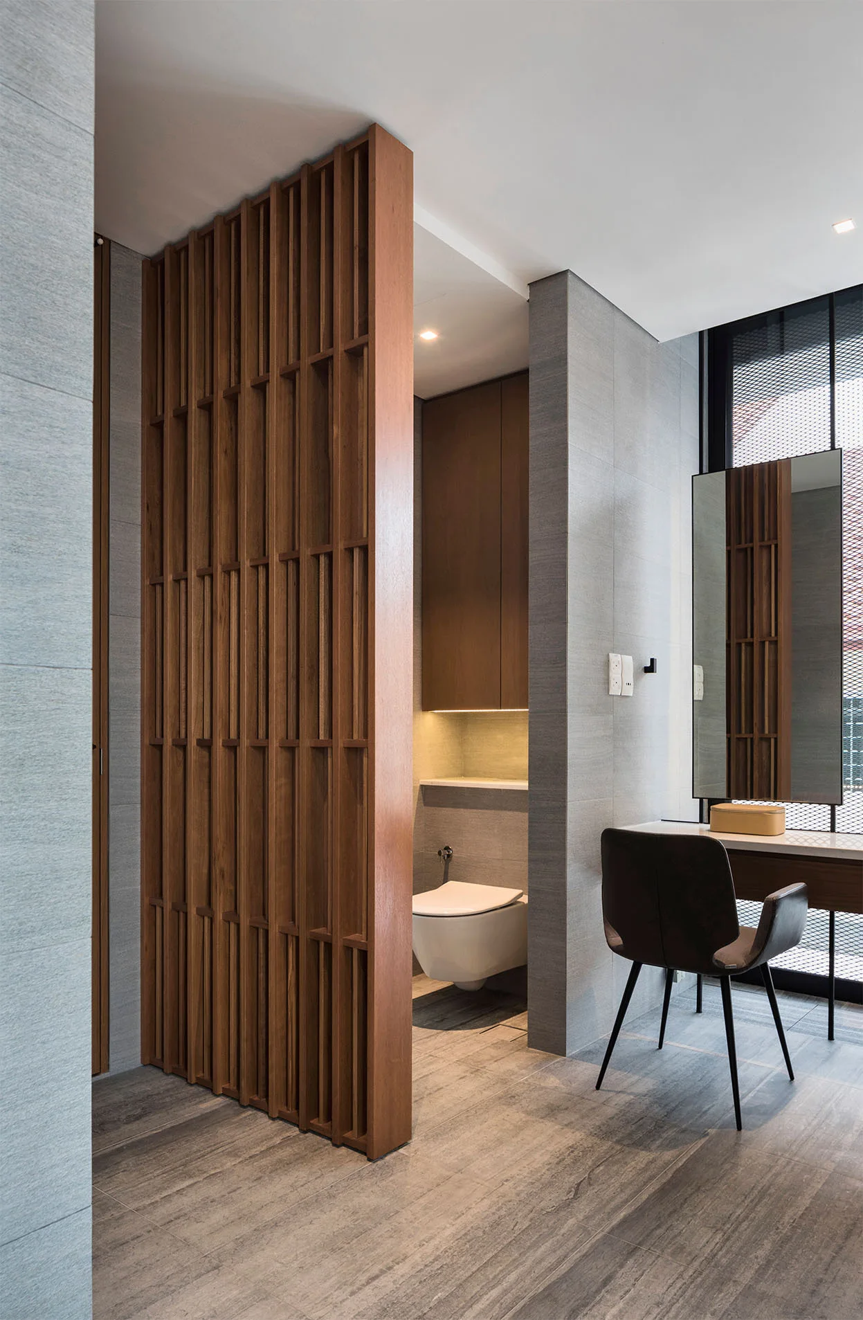 A modern bathroom with a grey tiled wall, a wall-mounted toilet, and a tall wood slat partition next to a vanity table.