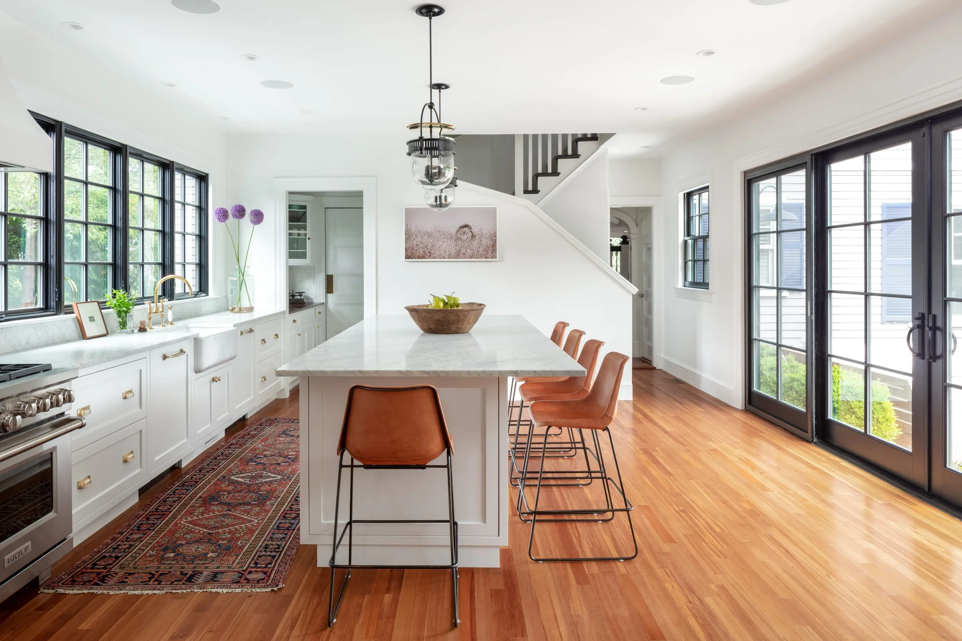A bright white kitchen with a large marble-topped island, four leather barstools, a patterned floor runner, and prominent black-framed windows.