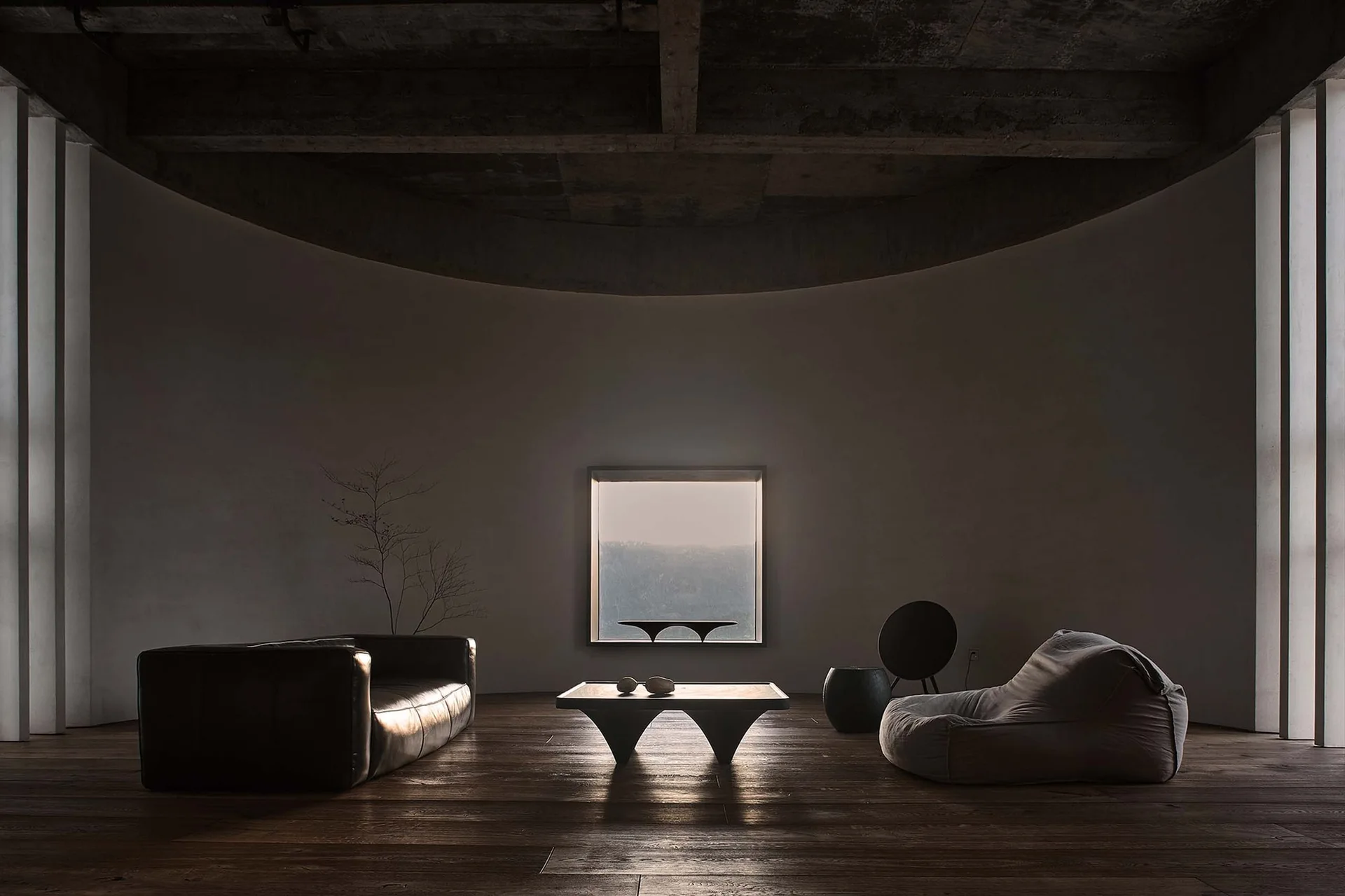 A minimalist living room with a curved concrete wall, a single square window, low-profile dark sofa, and a fabric beanbag on dark wood floors.