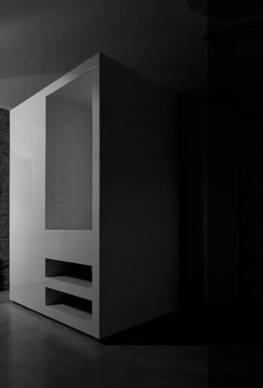 A minimalist partition block wrapped in glossy white square ceramic tiles features a tall recessed shower niche and two smaller lower shelves.