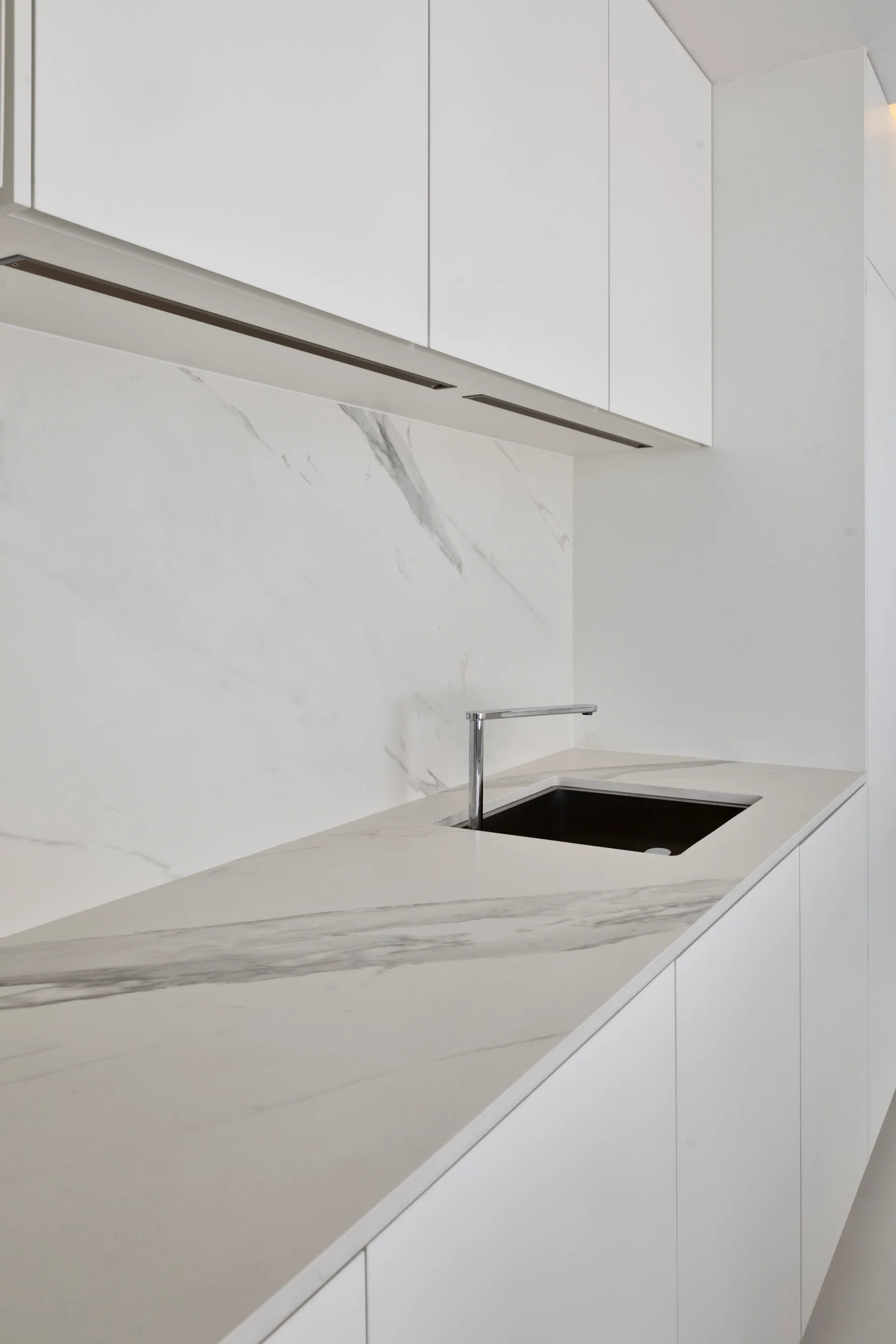 A close-up of a minimalist kitchen featuring flat-panel white cabinets, a veined marble countertop and backsplash, and a modern chrome faucet.
