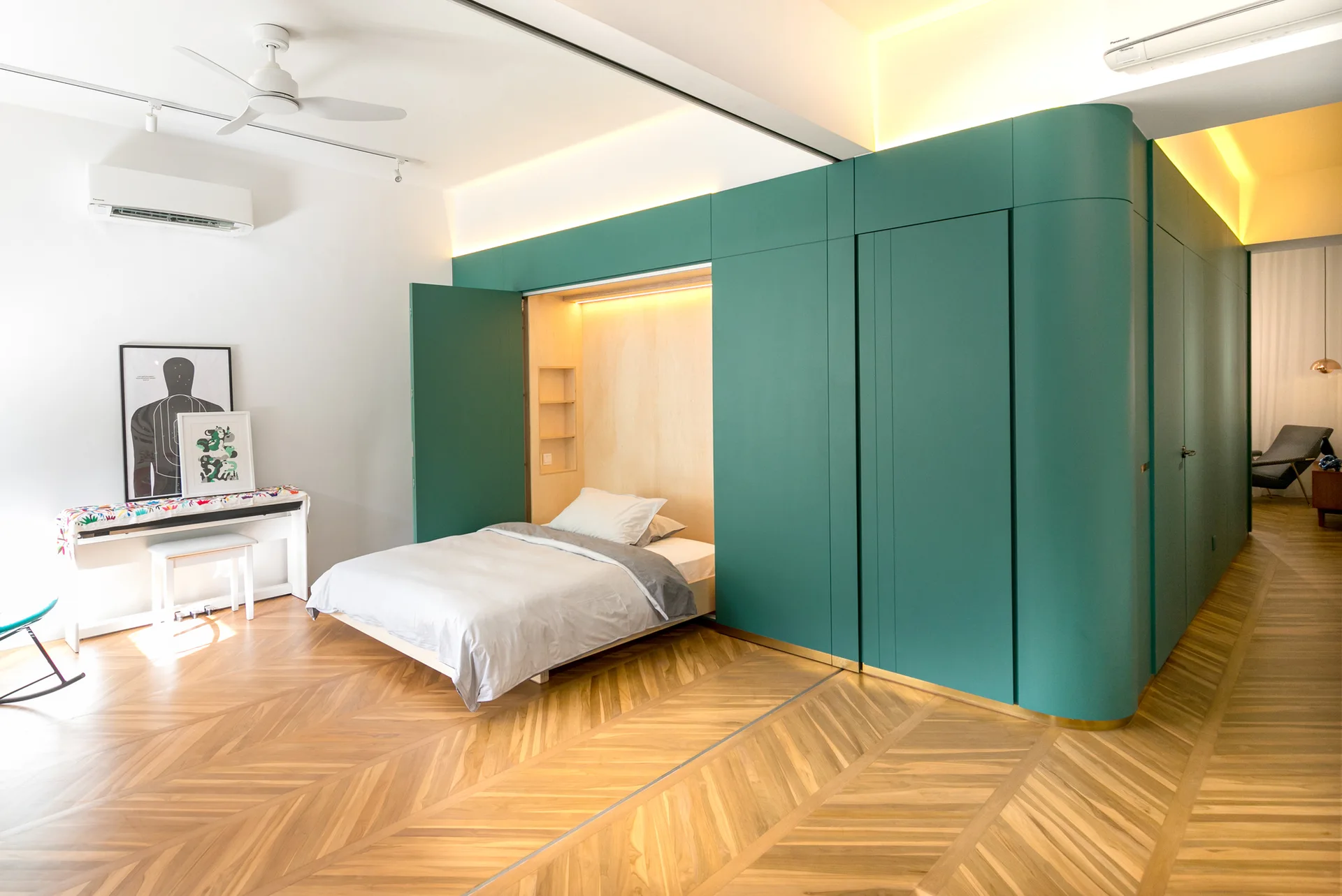 A modern bedroom features a large teal storage wall with an integrated fold-down bed open, set on herringbone wood flooring.