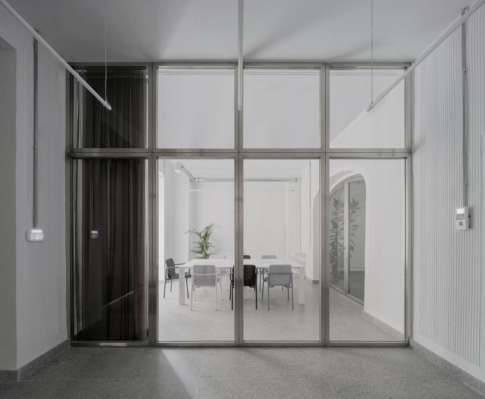 A minimalist conference room with a white table and chairs viewed through a large metal-framed glass partition with textured ribbed white walls.
