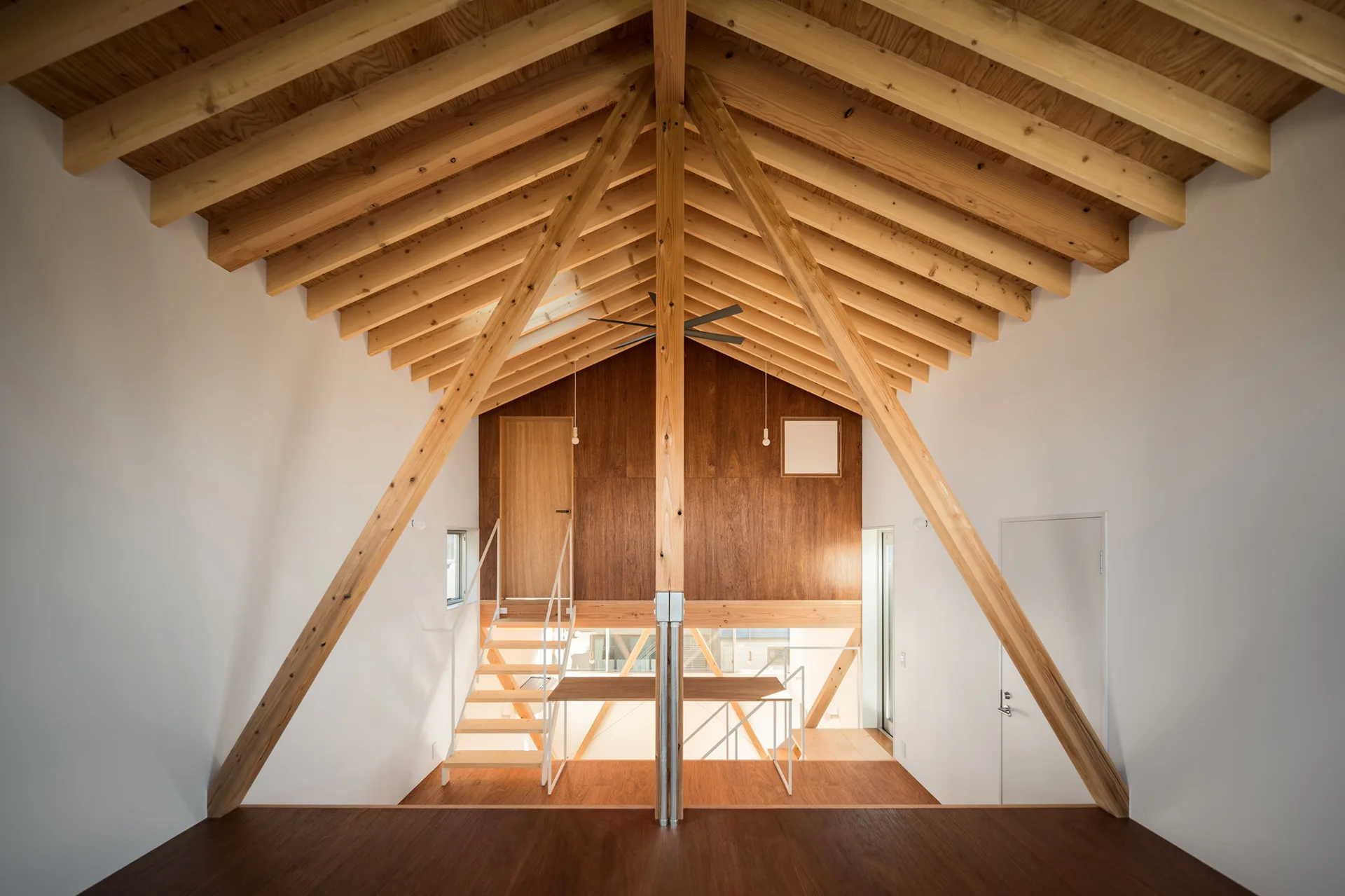 A symmetrical minimalist loft interior featuring exposed wooden roof trusses, white walls, and a wood-paneled upper-level wall with a staircase.