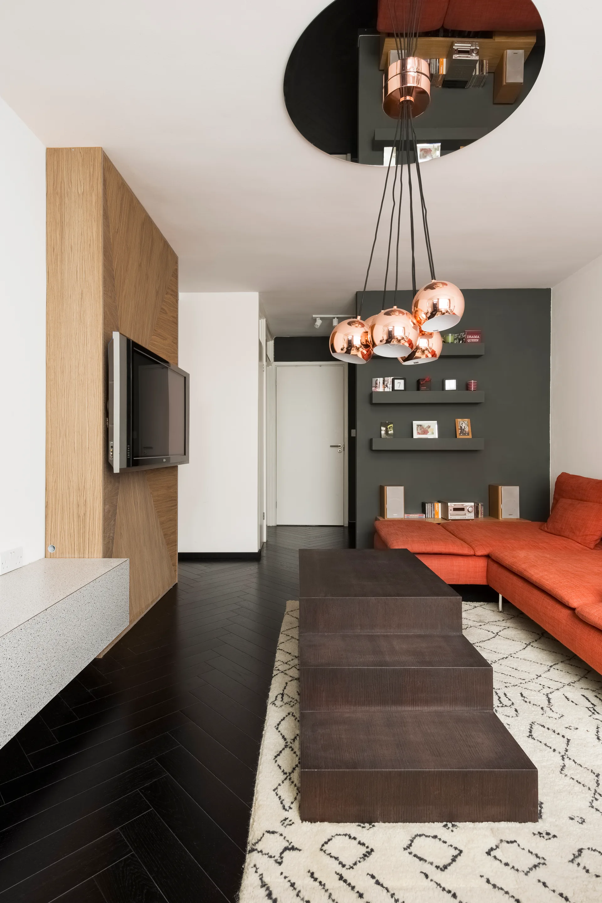 A modern living room features a bright orange sectional sofa, a stepped dark wood coffee table, and copper pendant lights over a patterned cream rug.