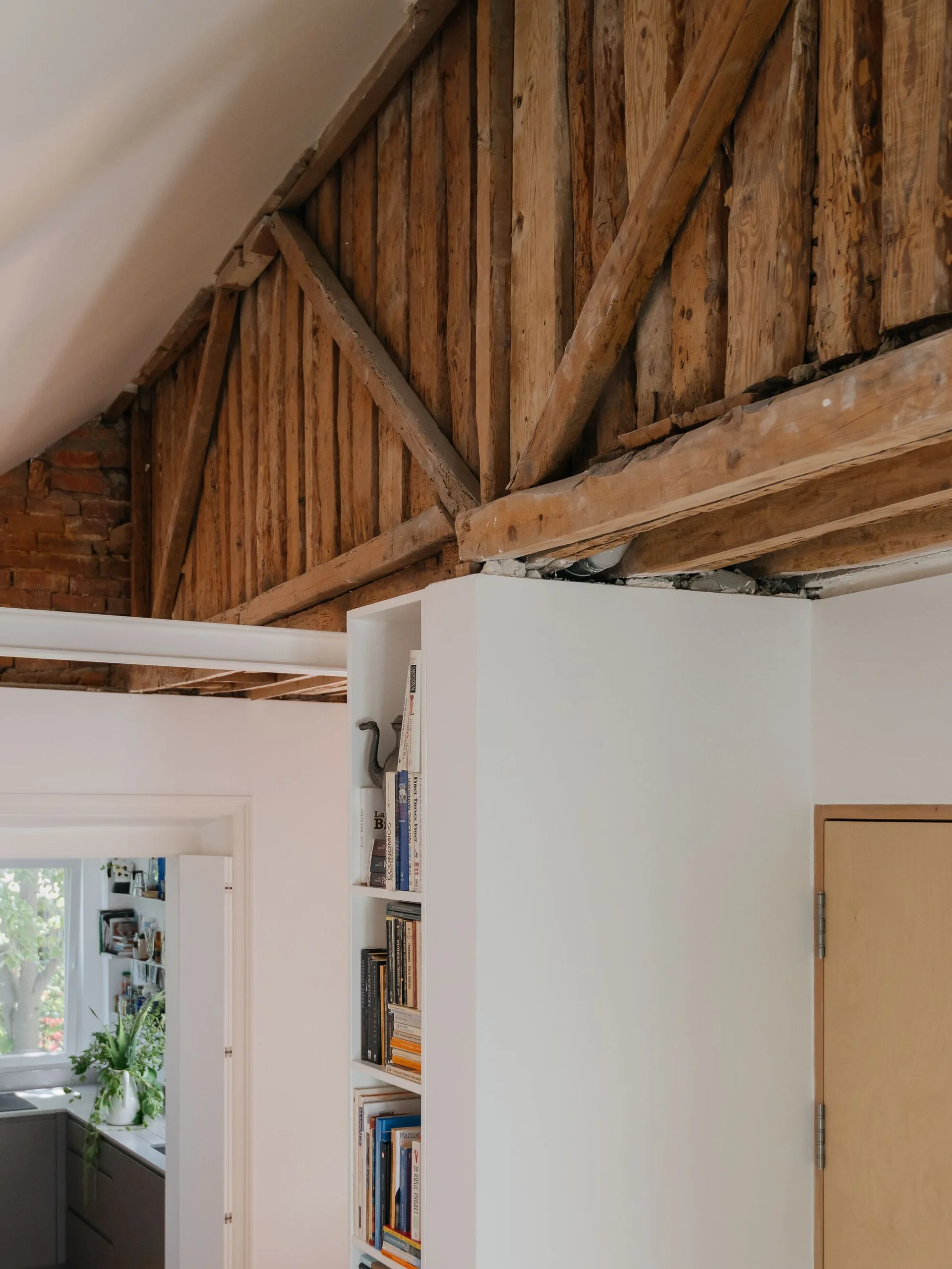 Raw structural timber trusses frame the ceiling of a room featuring a crisp white built-in bookshelf and smooth plaster walls.