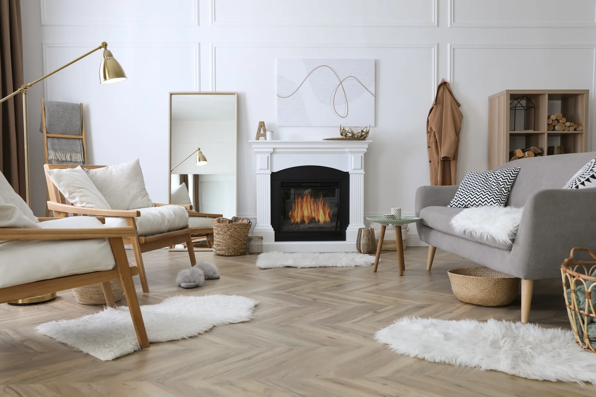 A light gray sofa, two wood-framed armchairs, and a white fireplace sit on a herringbone wood floor accented by small faux fur rugs.