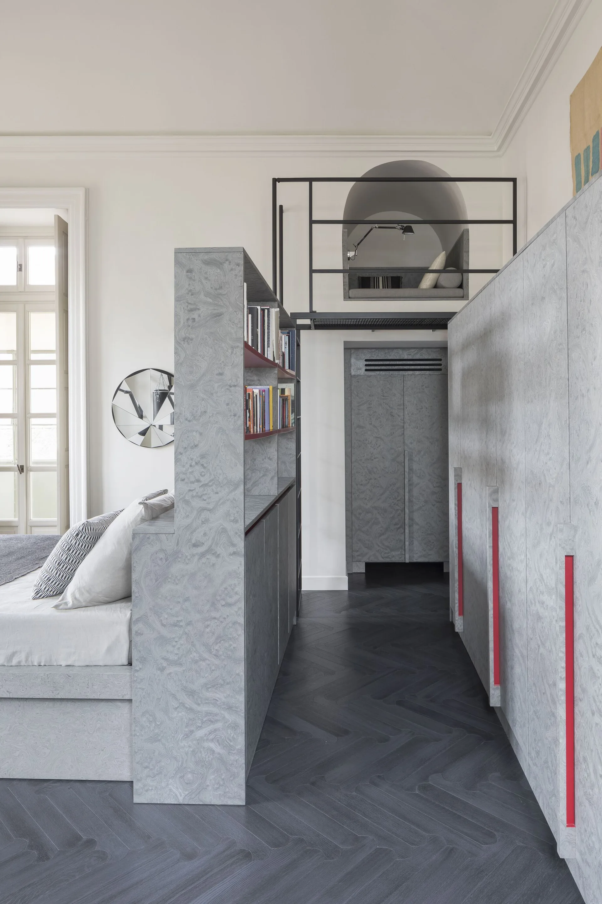 A modern bedroom featuring a grey burl wood bookshelf divider, matching wardrobe cabinets with red accents, and a small mezzanine loft.