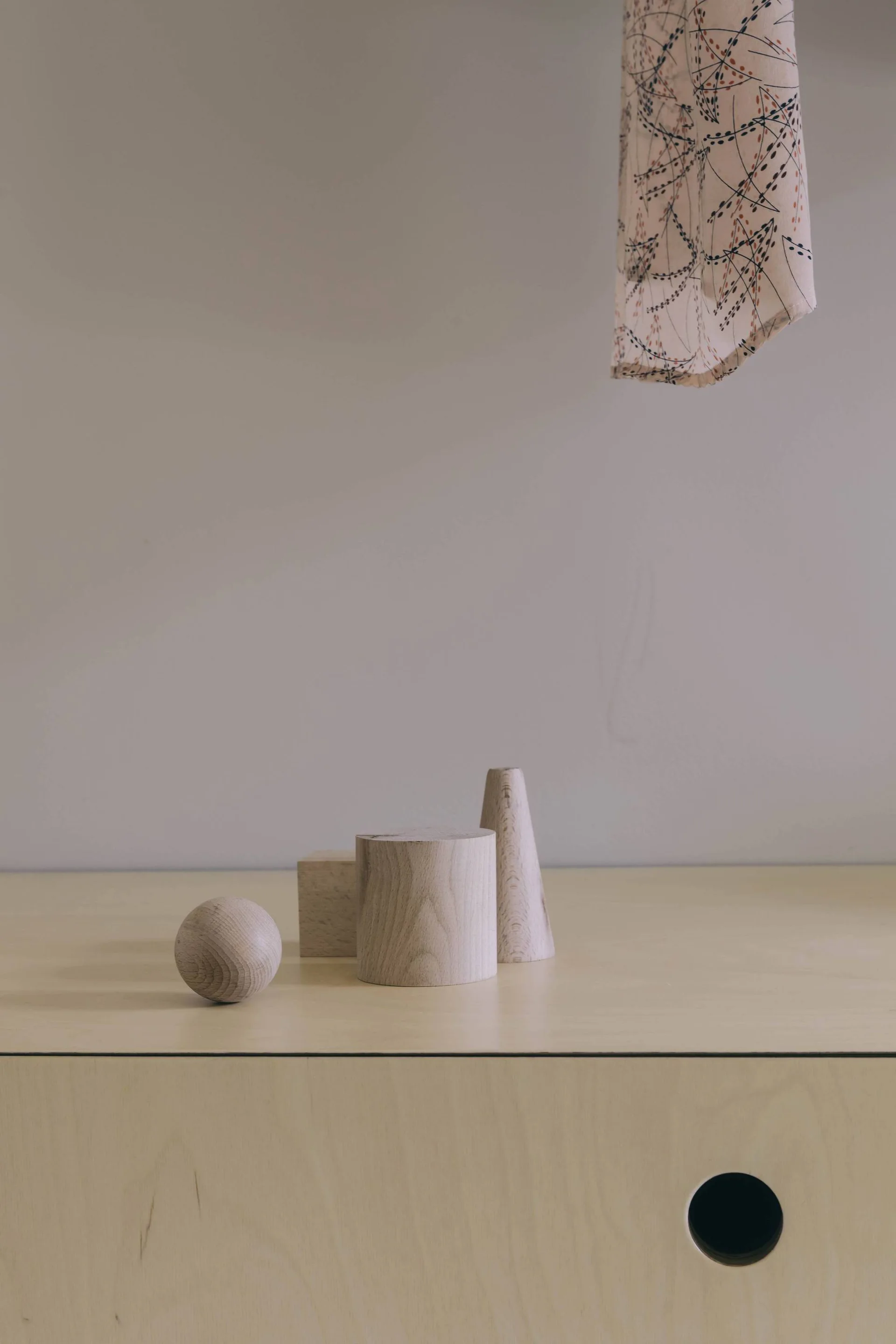 A minimalist arrangement of a wooden sphere, cube, cylinder, and cone on a light plywood cabinet against a smooth beige wall.